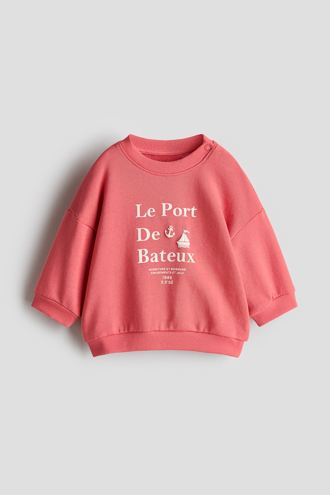 Un sweat-shirt rose est présenté de face, muni d'un col ras-du-cou côtelé avec deux boutons-pression sur l'épaule gauche. L'avant porte un texte blanc "Le Port De Bateaux" accompagné d'une ancre et d'une icône de voilier, et est fini par des poignets et un bas côtelés. - 1