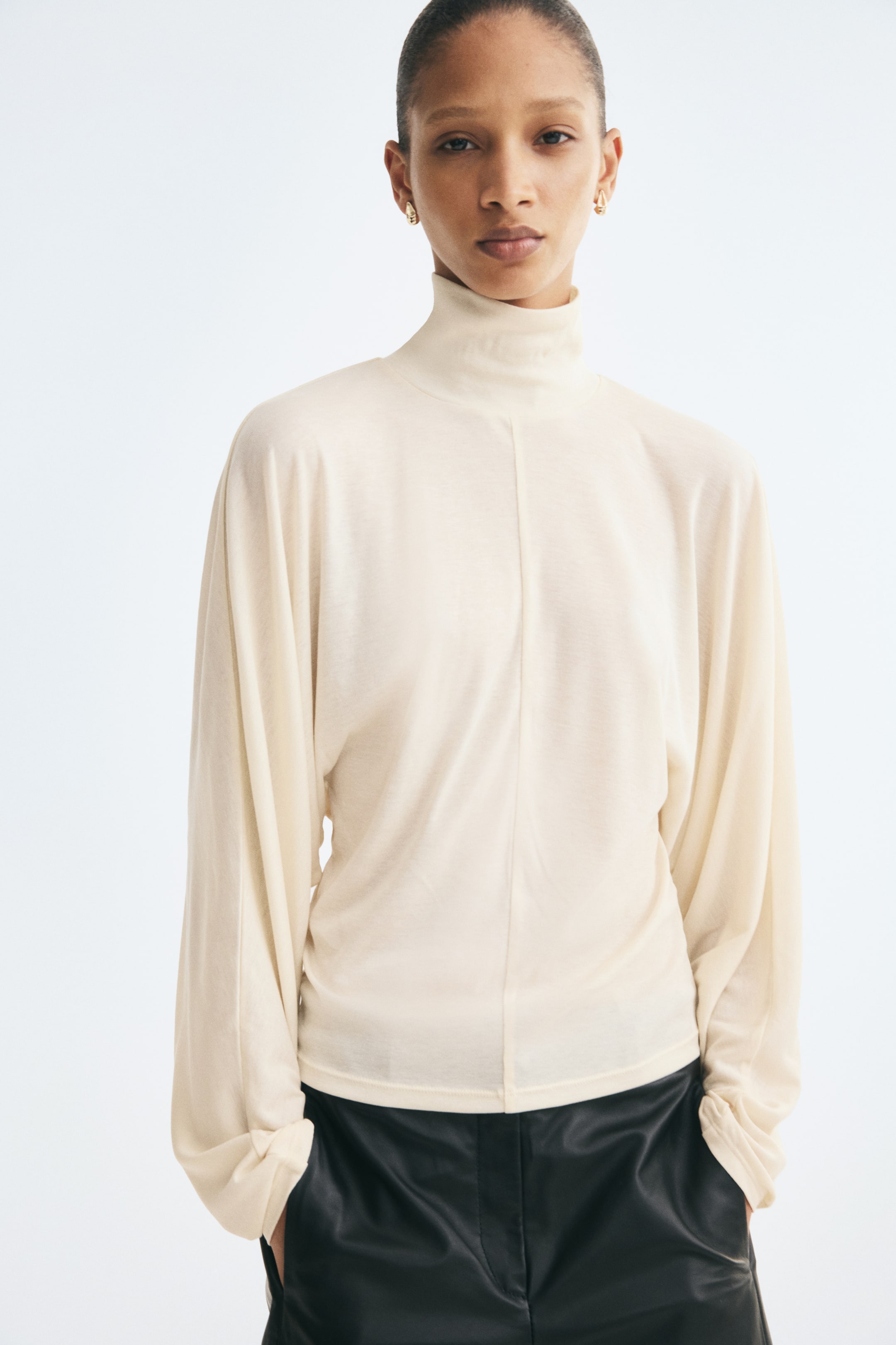 Ampliar la imagen: Se muestra de frente un top de punto fino color crema, con cuello alto de doble capa, mangas batwing largas y drapeadas, y una costura vertical prominente que recorre el centro. El top se combina con pantalones negros, resaltando su tejido suave y su silueta relajada pero ajustada.