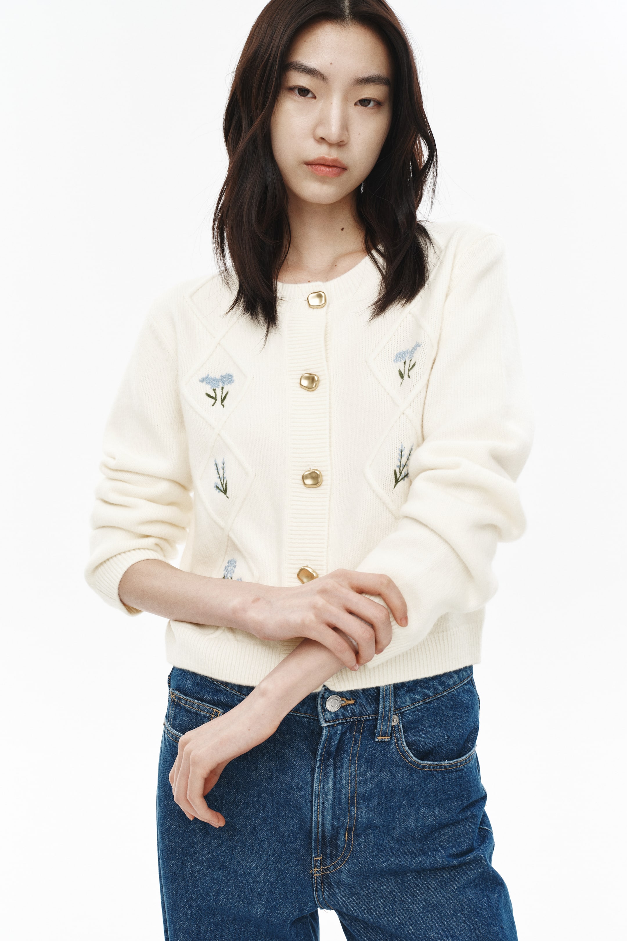 이미지 확대 보기: 크림색 카디건은 라운드 네크라인과 다이아몬드 니트 패턴에 파란색 꽃 자수가 특징입니다. 금색 톤의 원형 버튼이 앞면 플래킷을 잠그고 있으며, 리브드 커프스와 리브드 밑단이 의상을 완성합니다.