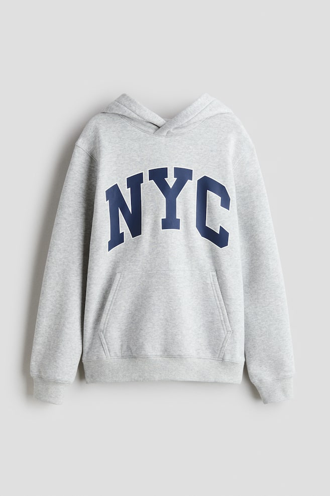 En lys grå hettegenser i bomullssweatshirtstoff blir presentert fra forsiden, med en foret hette med omslag foran, droppede skuldre og store, mørkeblå blokkbokstaver som staver 'NYC' på brystet. En kengurulomme er synlig under teksten, med ribbestrikkede mansjetter som avslutter de lange ermene og en ribbestrikket kant som fullfører nederkanten. - 1