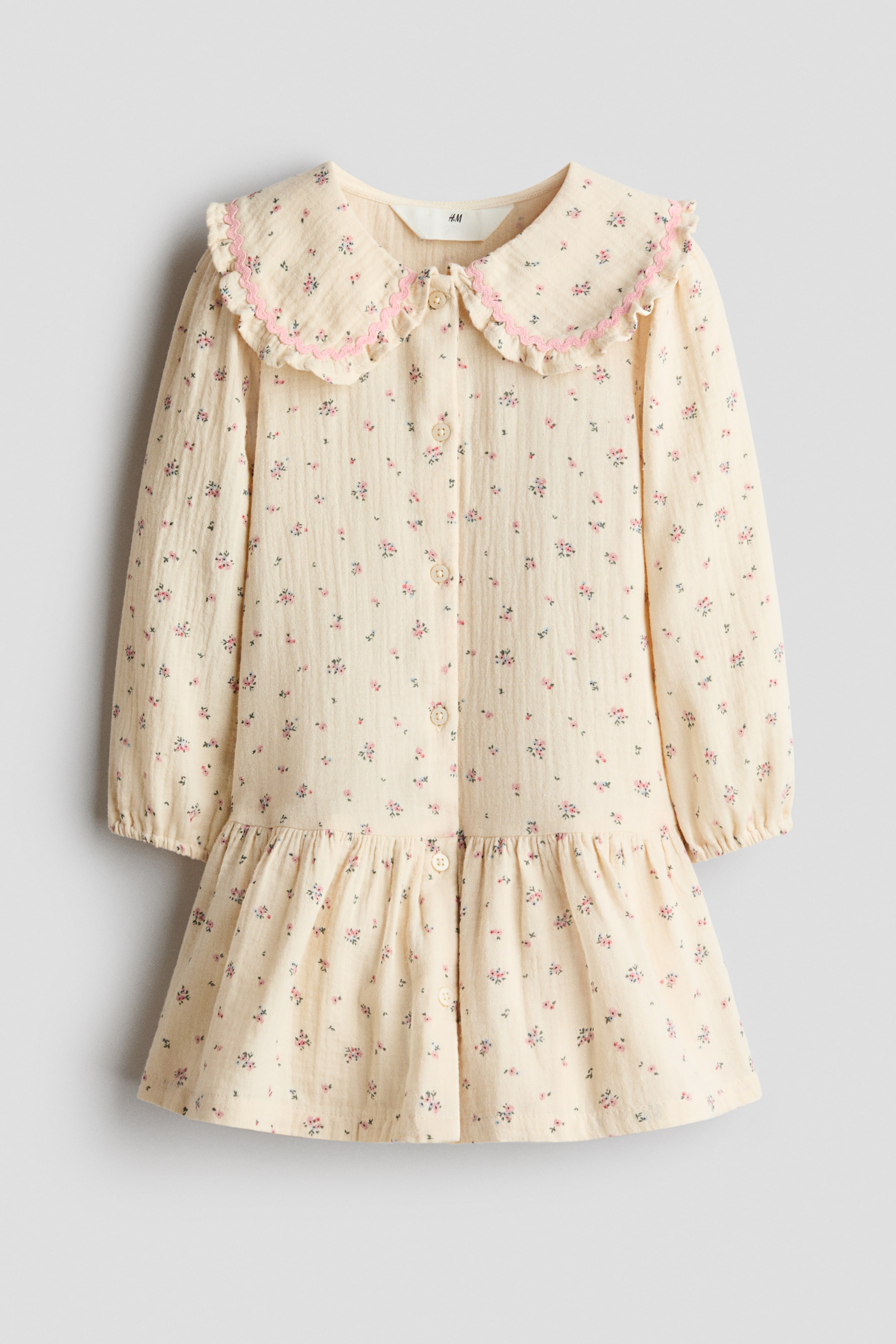 View larger image: A cream textured dress with a small pink floral print is shown from the front, featuring a rounded Peter Pan collar with pink ruffled trim, long sleeves with elasticated cuffs, a full button-down front with cream buttons, and a gathered tiered skirt.