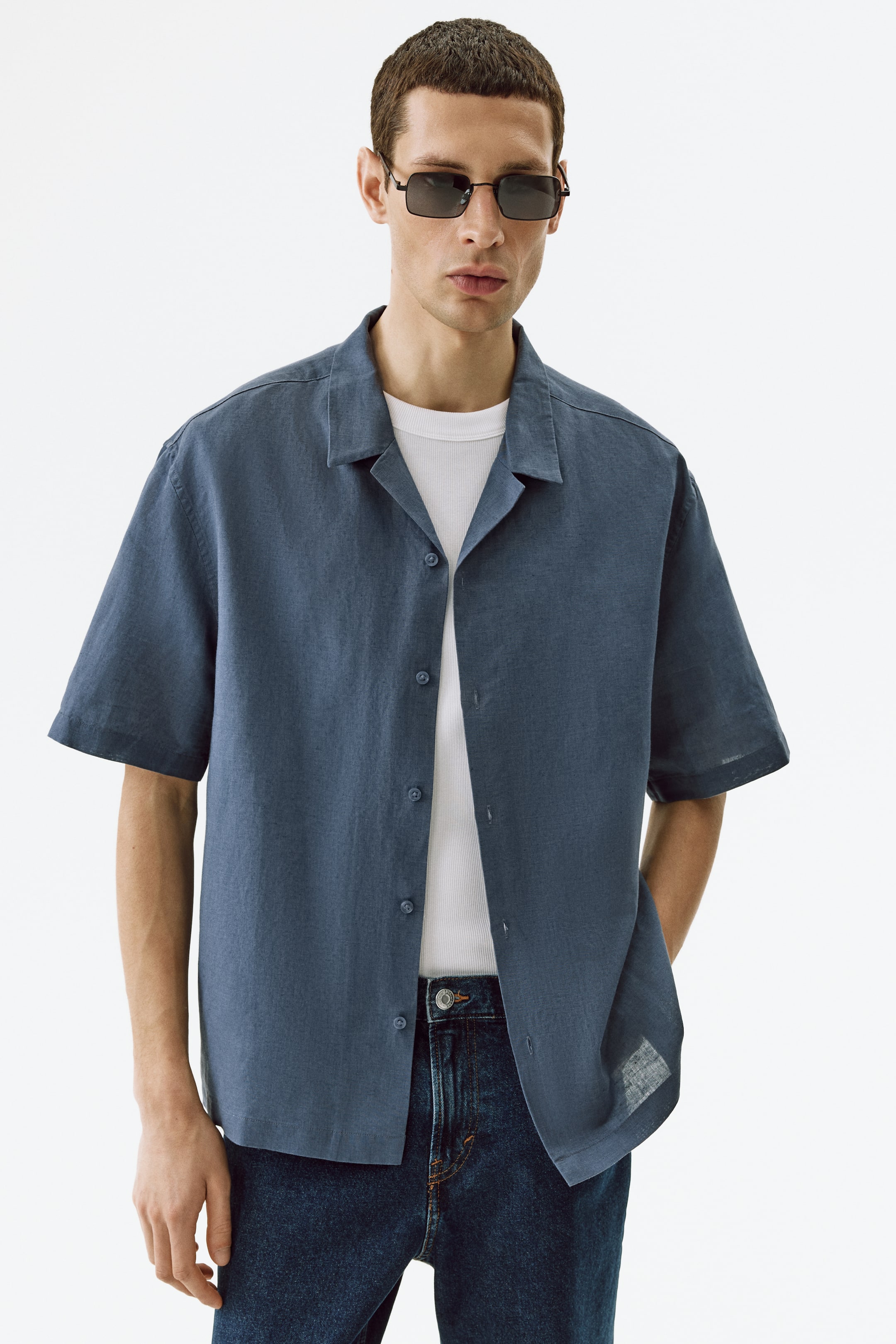 Visualizza immagine più grande: Una camicia blu a maniche corte presenta un colletto alla cubana e una chiusura frontale con bottoni, indossata aperta su una t-shirt bianca a coste. La camicia a vestibilità rilassata mette in mostra un tessuto strutturato, con l'orlo dritto che poggia su jeans scuri lavaggio.