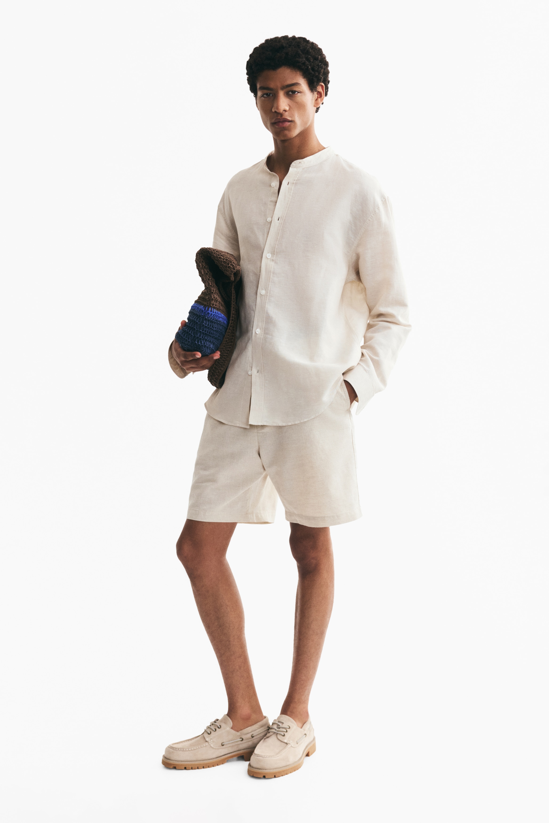 Light beige shorts are presenting a front view, laid flat, featuring an elasticated waistband with a drawstring and a button closure. Side seam pockets are visible, and the fabric is showing a textured cotton and linen weave.