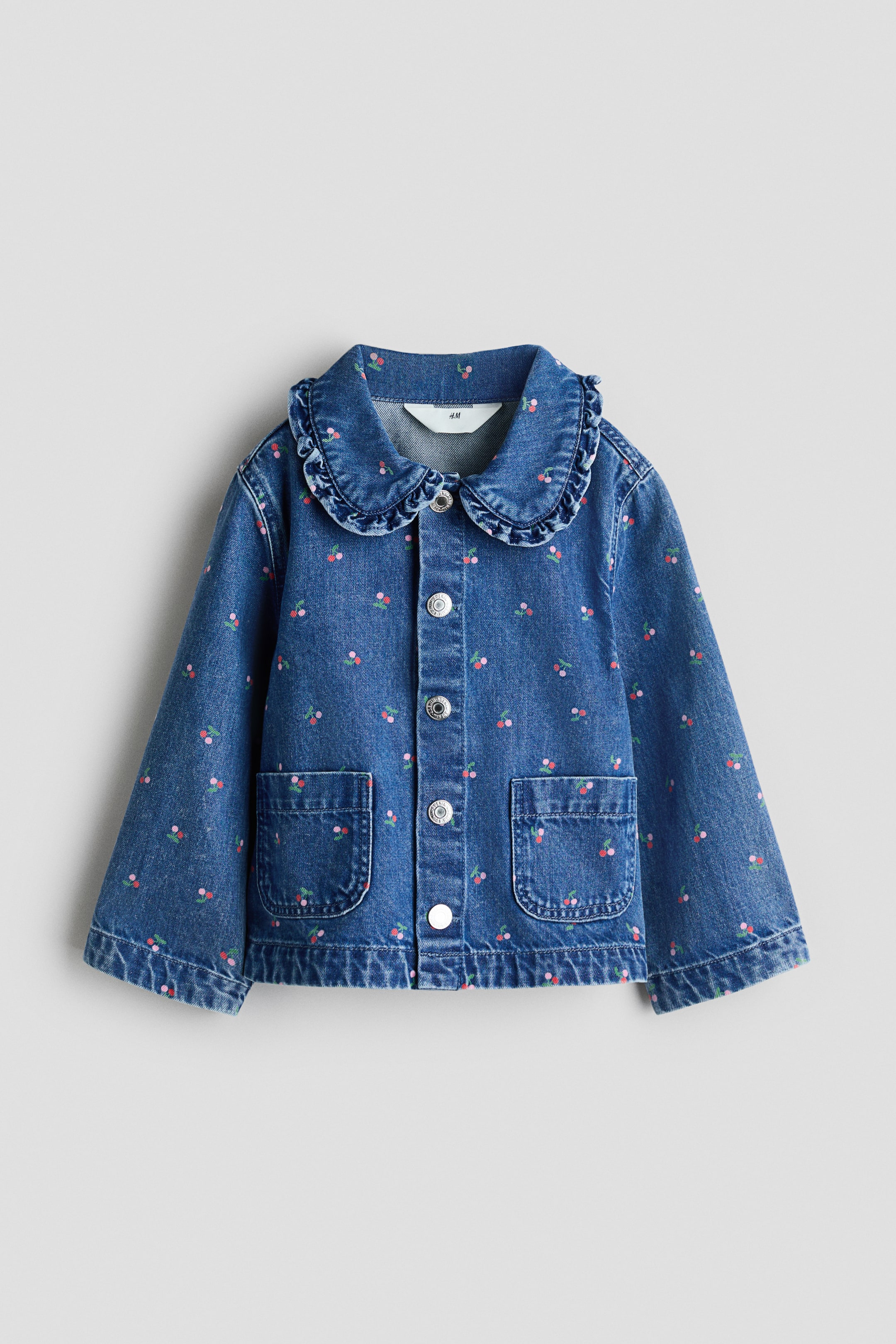 View larger image: A blue denim jacket is showcasing its front, adorned with an all-over small cherry print. It is crafted with a ruffled Peter Pan collar, a front button closure with silver-toned buttons, and two patch pockets on the front.