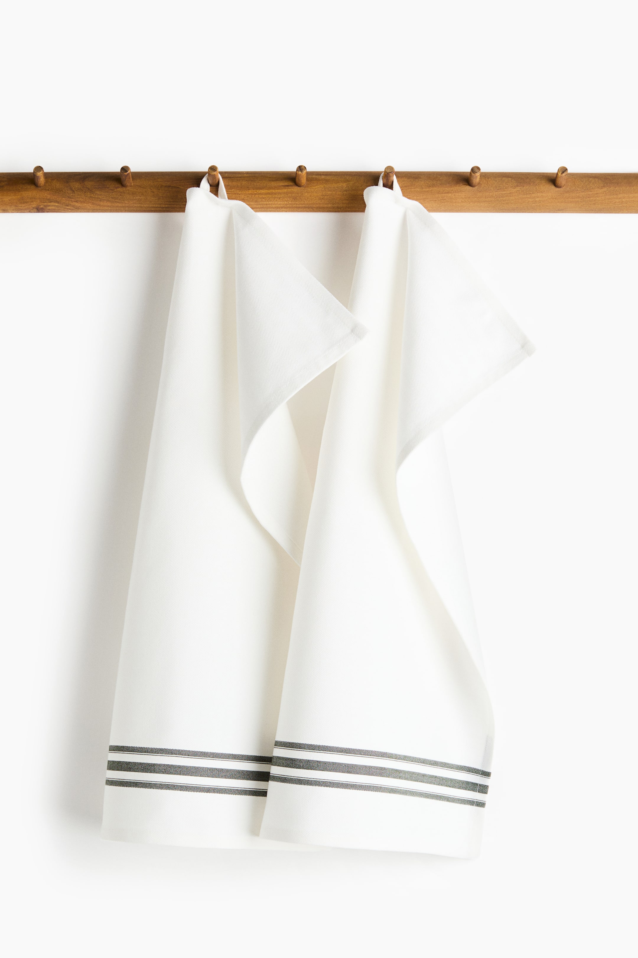 View larger image: Two white cotton tea towels featuring a herringbone weave are hanging from their fabric loops on a wooden peg rail. The towels are draping downwards, showing horizontal, dark grey yarn-dyed stripes running above the bottom hems.