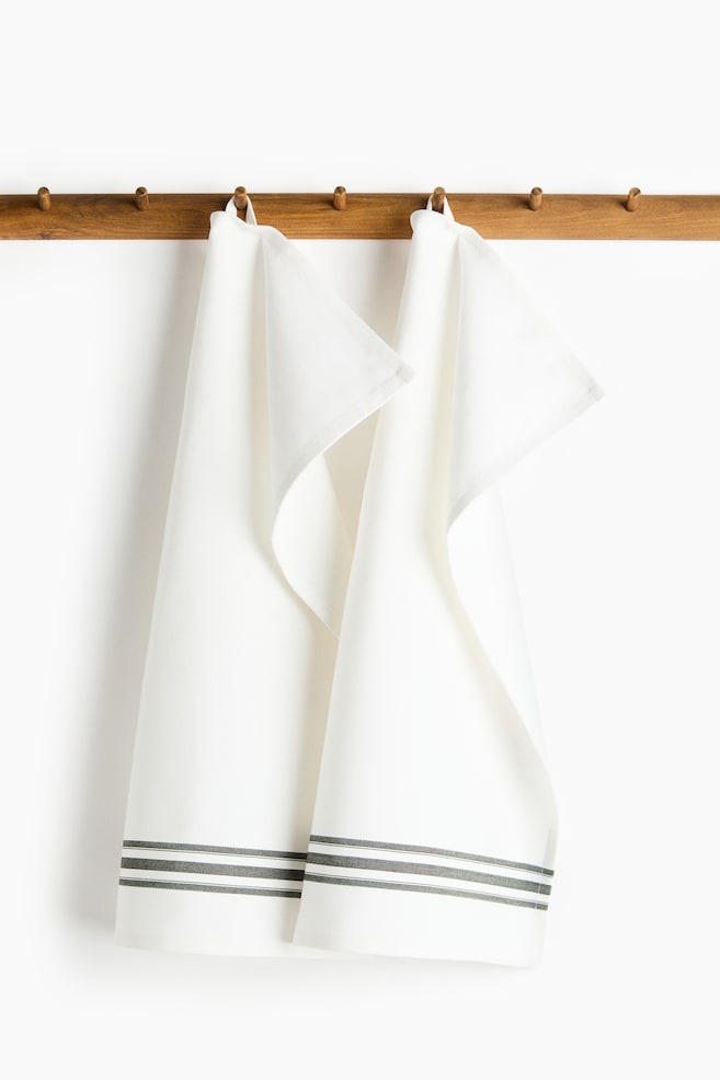 Two white cotton tea towels featuring a herringbone weave are hanging from their fabric loops on a wooden peg rail. The tea towels are draping downwards, showing horizontal, dark grey yarn-dyed stripes running above the bottom hems. - 1