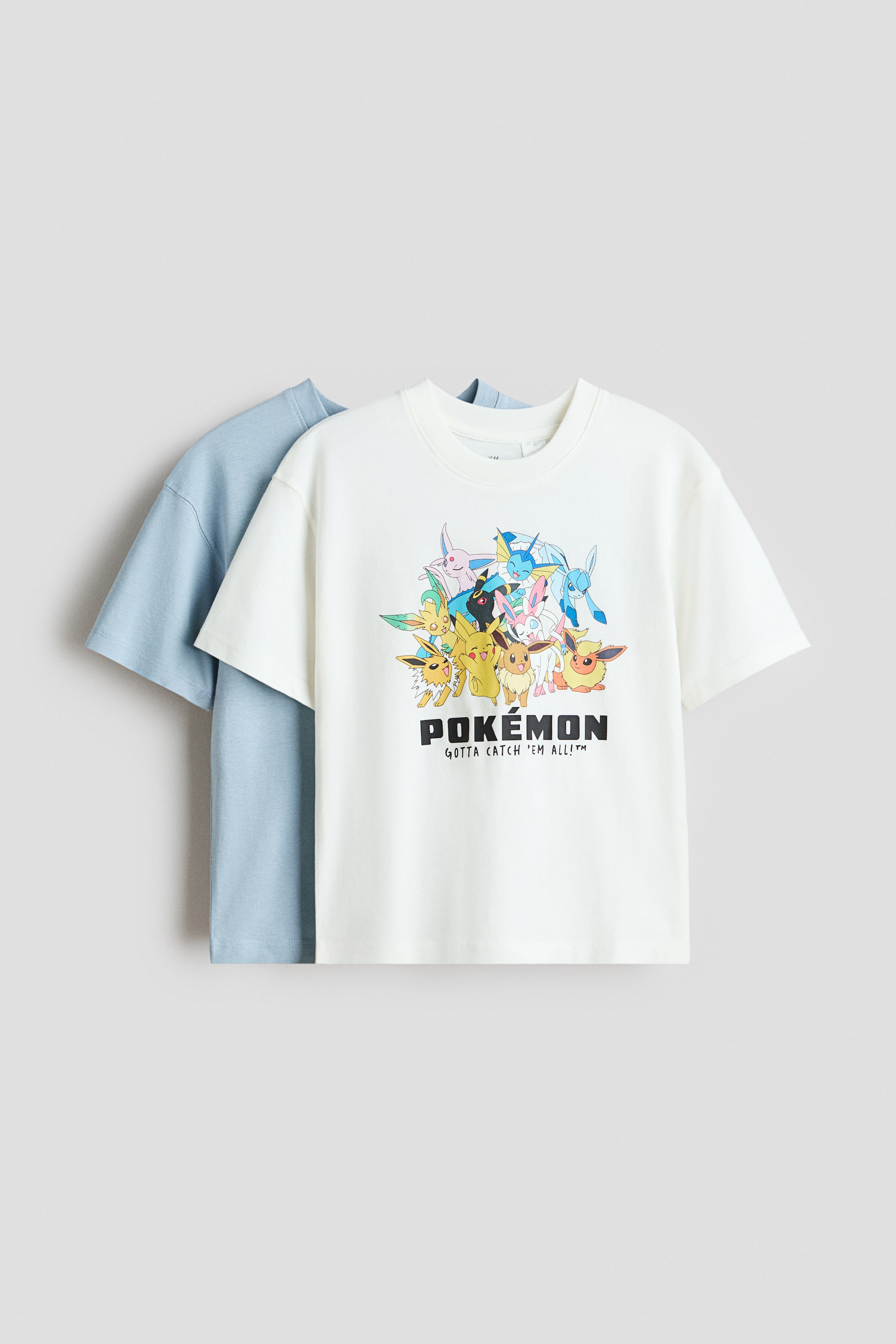 Xem ảnh lớn hơn: Hai chiếc áo phông dáng rộng bằng vải cotton jersey mềm mại đang được trình bày với góc nhìn từ phía trước. Chiếc áo phông trắng nổi bật với hình in nhân vật Pok�mon đầy màu sắc và chữ, cổ áo tròn viền gân, và vai rớt, với một chiếc áo phông màu xanh nhạt lộ ra một phần phía sau.
