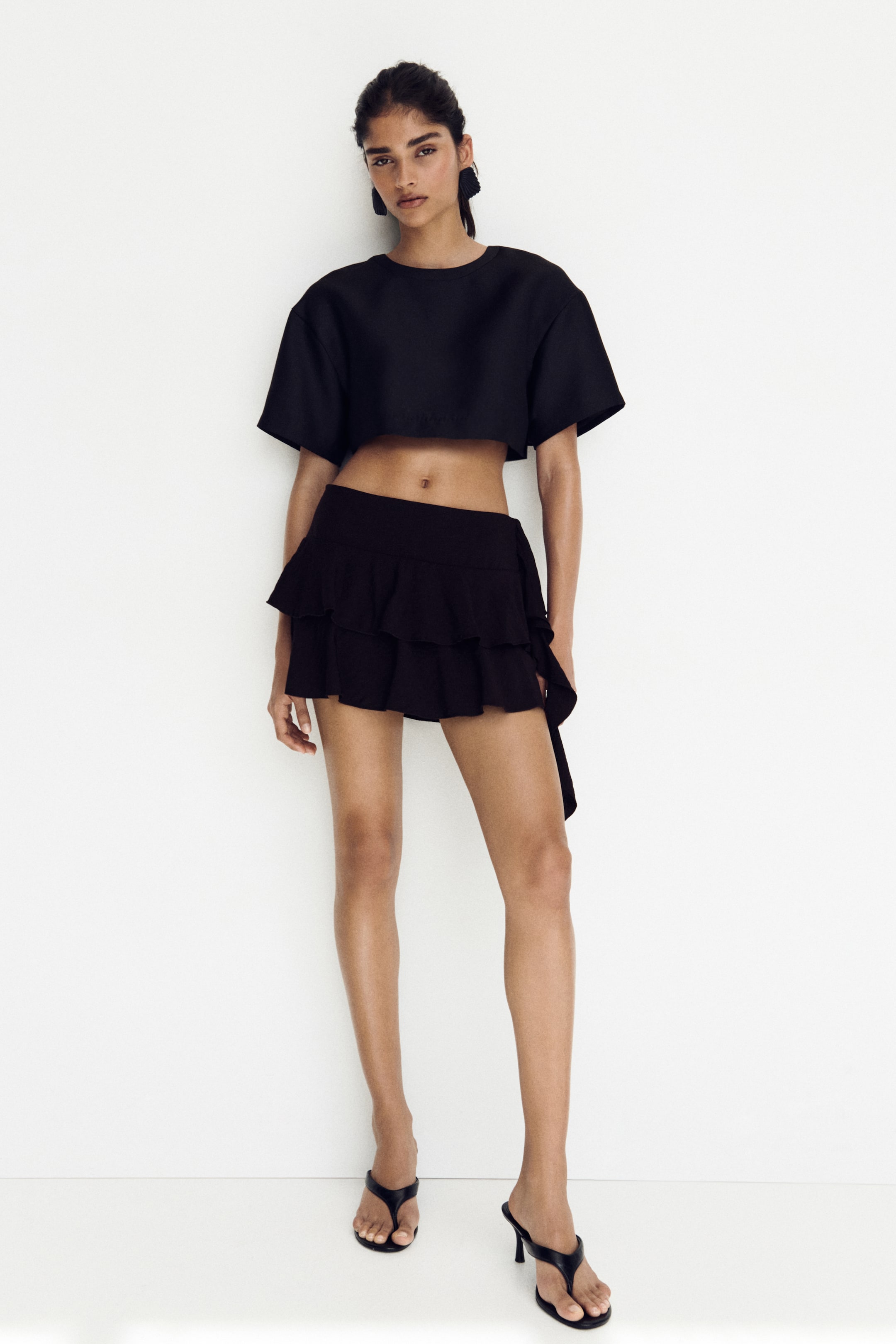 View larger image: A black short-sleeved cropped top features a wide, boxy cut with a crew neck and an elasticized hem, paired with a black ruffled mini skirt showcasing two tiers of ruffles and a wrap-style tie detail.
