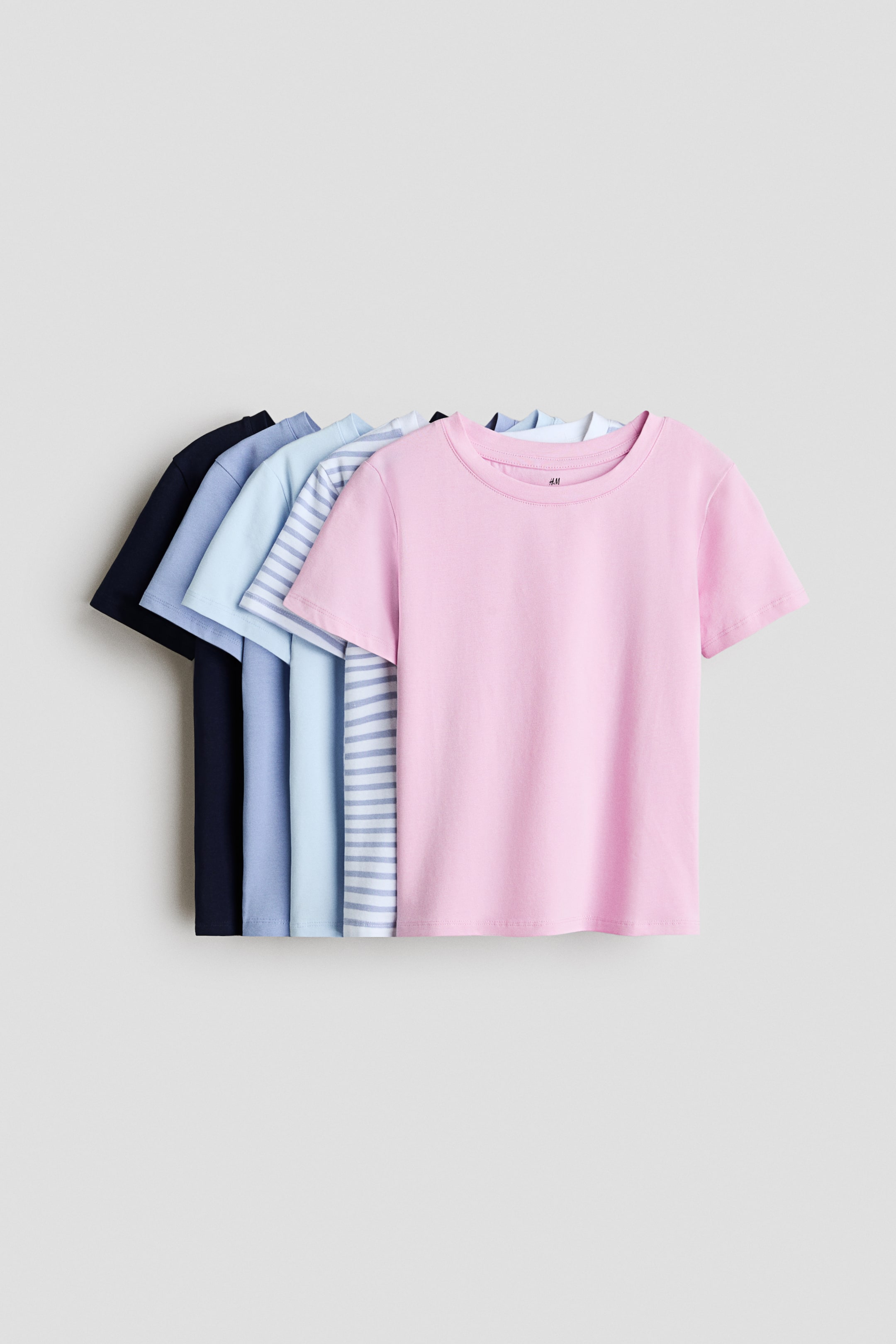 View larger image: Five short-sleeved crewneck T-shirts are arranged in an overlapping row facing front, displaying a color gradient from dark navy blue at the back to solid light pink at the front. The middle layers feature solid shades of medium blue and light blue, along with a white shirt decorated with light blue horizontal stripes.