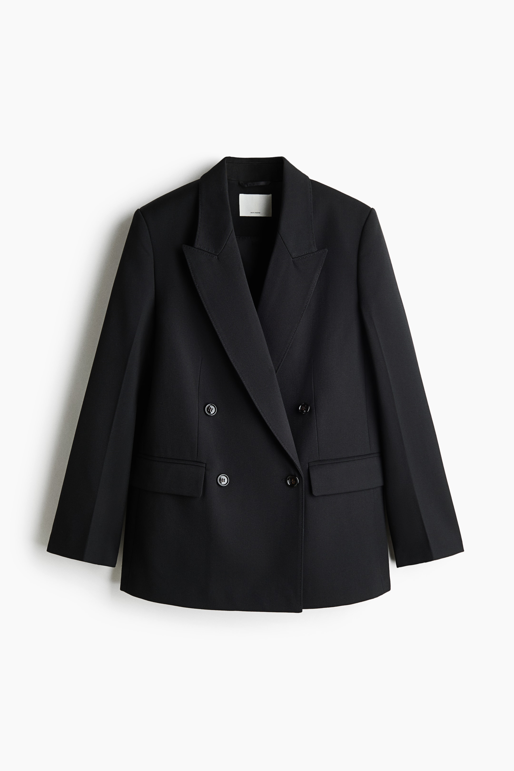 View larger image: A long black double-breasted blazer is hanging front-facing, featuring wide peak lapels and a four-button closure. The blazer showcases structured shoulders, front waist darts, and two jetted pockets with flaps.