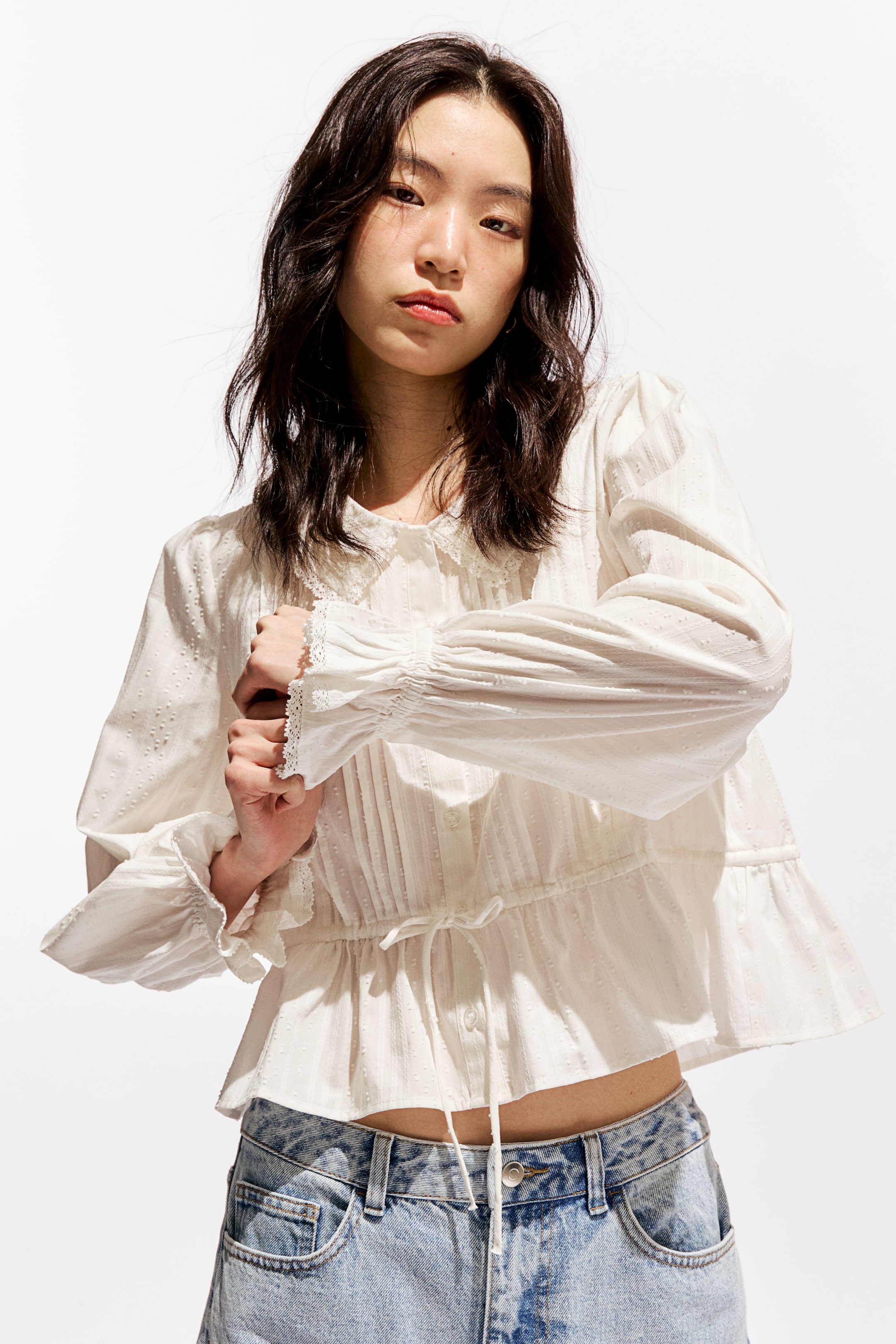 拡大画像を表示: 白いテクスチャードブラウスは、丸いレーストリム付きの襟、部分的なボタンプラケット、そしてボディスに縦のピンタックが特徴です。長いボリュームのある袖には、ギャザーされたレーストリム付きのカフスがあり、ウエストを絞るドローストリングが施されています。裾にはフリルのペプラムがあしらわれています。