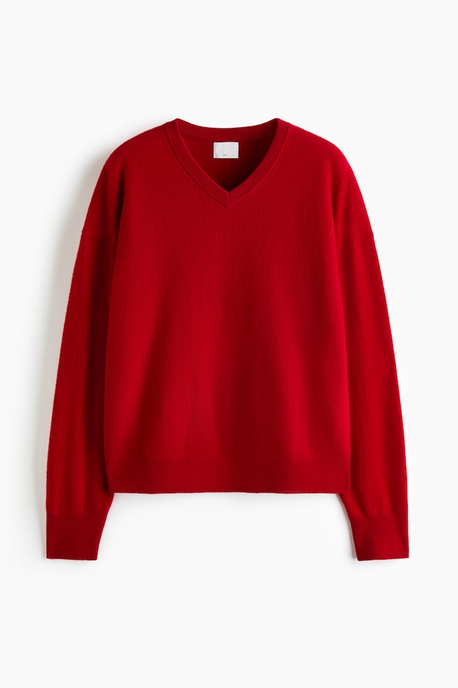 An oversized red wool jumper is facing forwards, displaying a fine-knit texture with a ribbed V-neck and dropped shoulders. The jumper features long sleeves with ribbed cuffs and a ribbed hem. - 1
