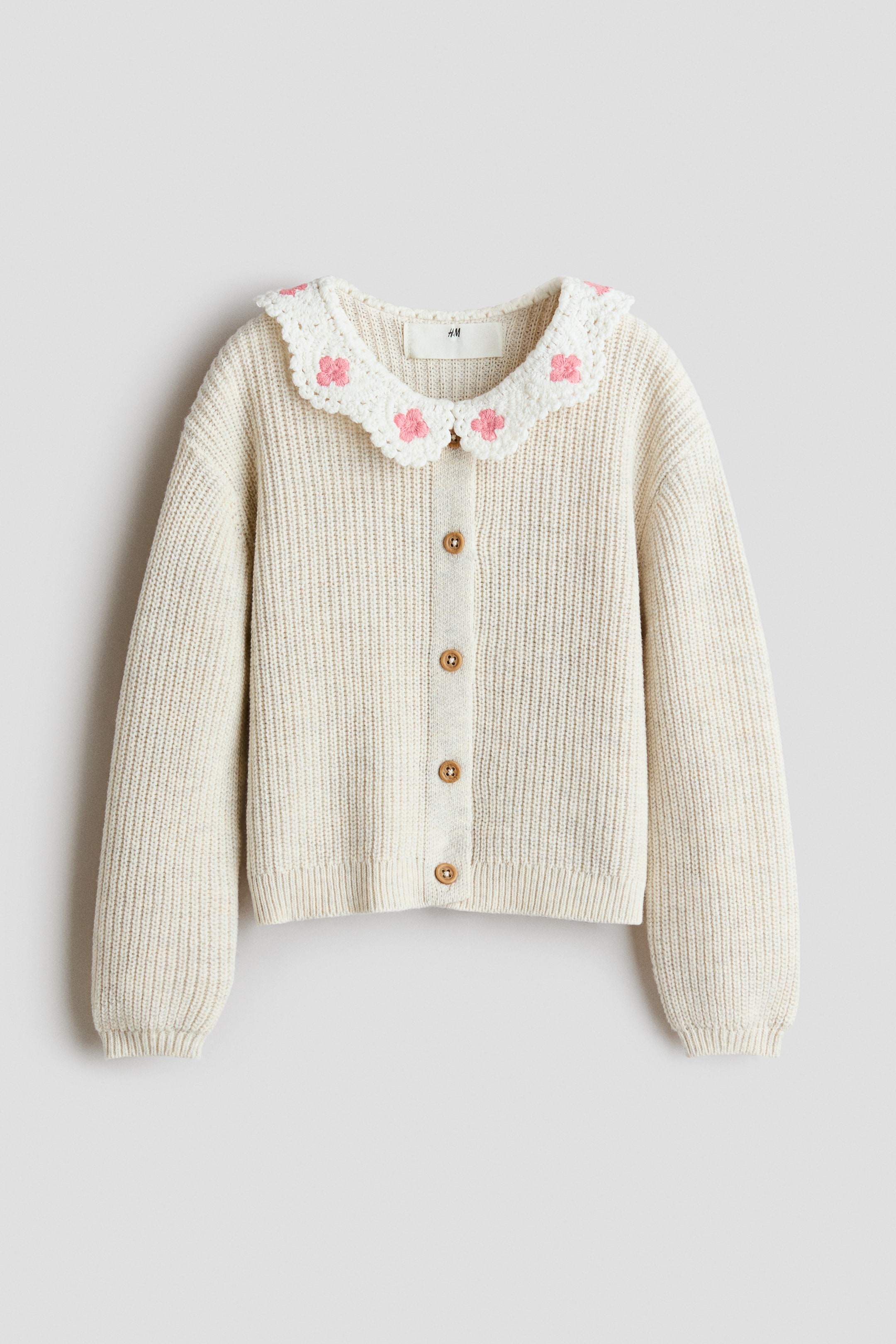 View larger image: A cream ribbed knit cardigan is showcasing a white crochet collar with pink floral motifs. It features five brown circular buttons down the front, along with ribbed cuffs and a ribbed hem.