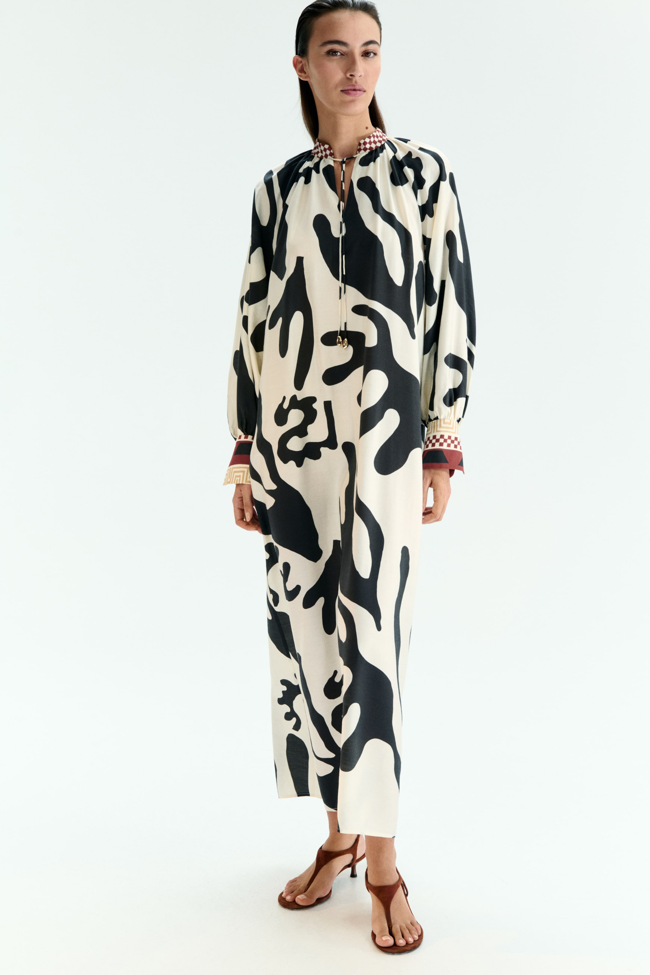 View larger image: A full-length dress, featuring a cream base with abstract black patterns, is being worn by the model, who is also wearing brown strappy sandals. The dress is showcasing a stand collar with a small checkerboard pattern, a V-neckline with a drawstring closure, and long sleeves ending in gathered cuffs with contrasting geometric patterns.
