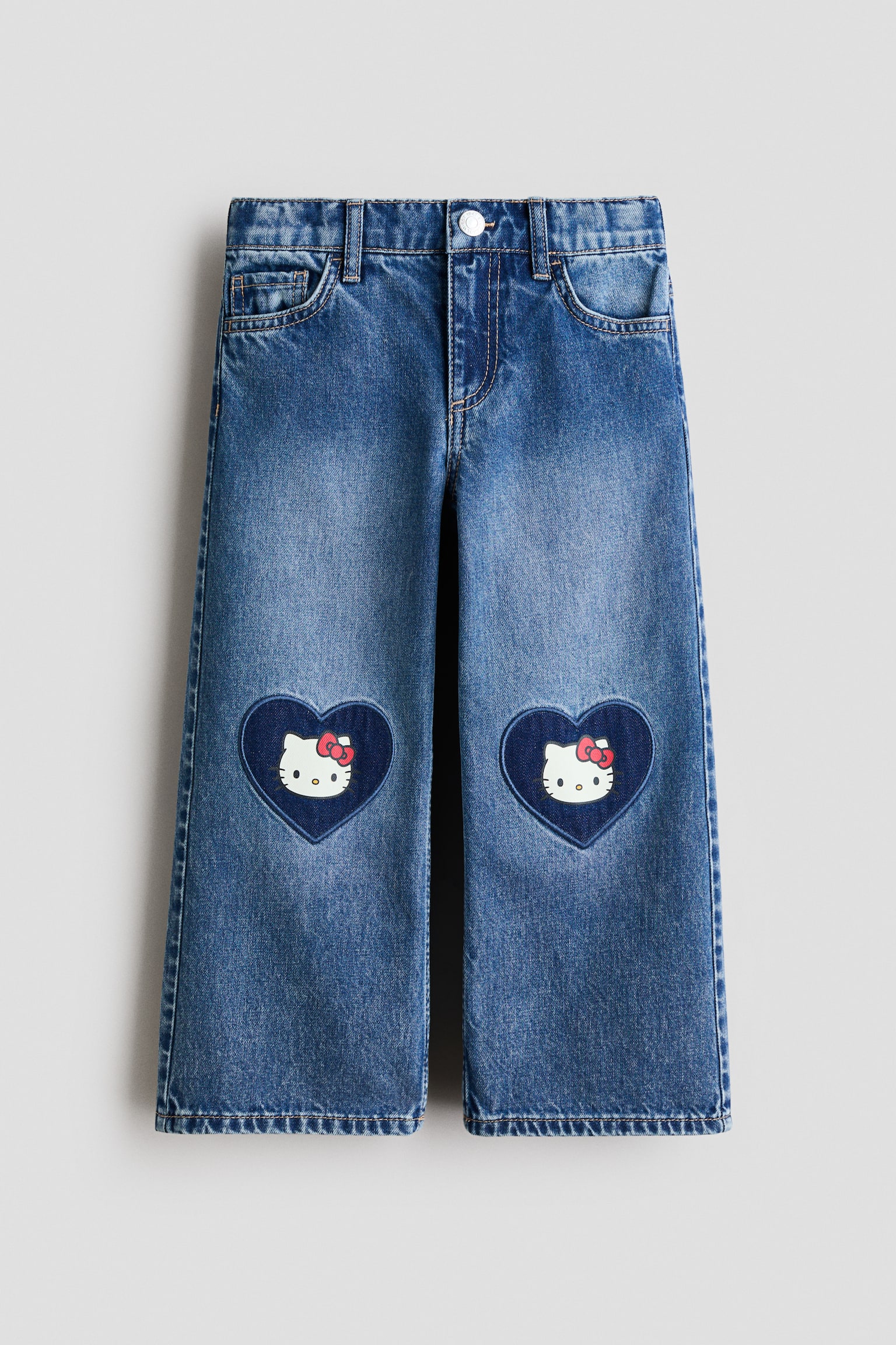 5-lomme jeans vises fra fronten, med en løs pasform gennem hofte og lår samt brede ben. Det blå denim fremviser hjerteformede Hello Kitty-patches på knæene, en lynlåsgylp og en trykknaplukning. - 1