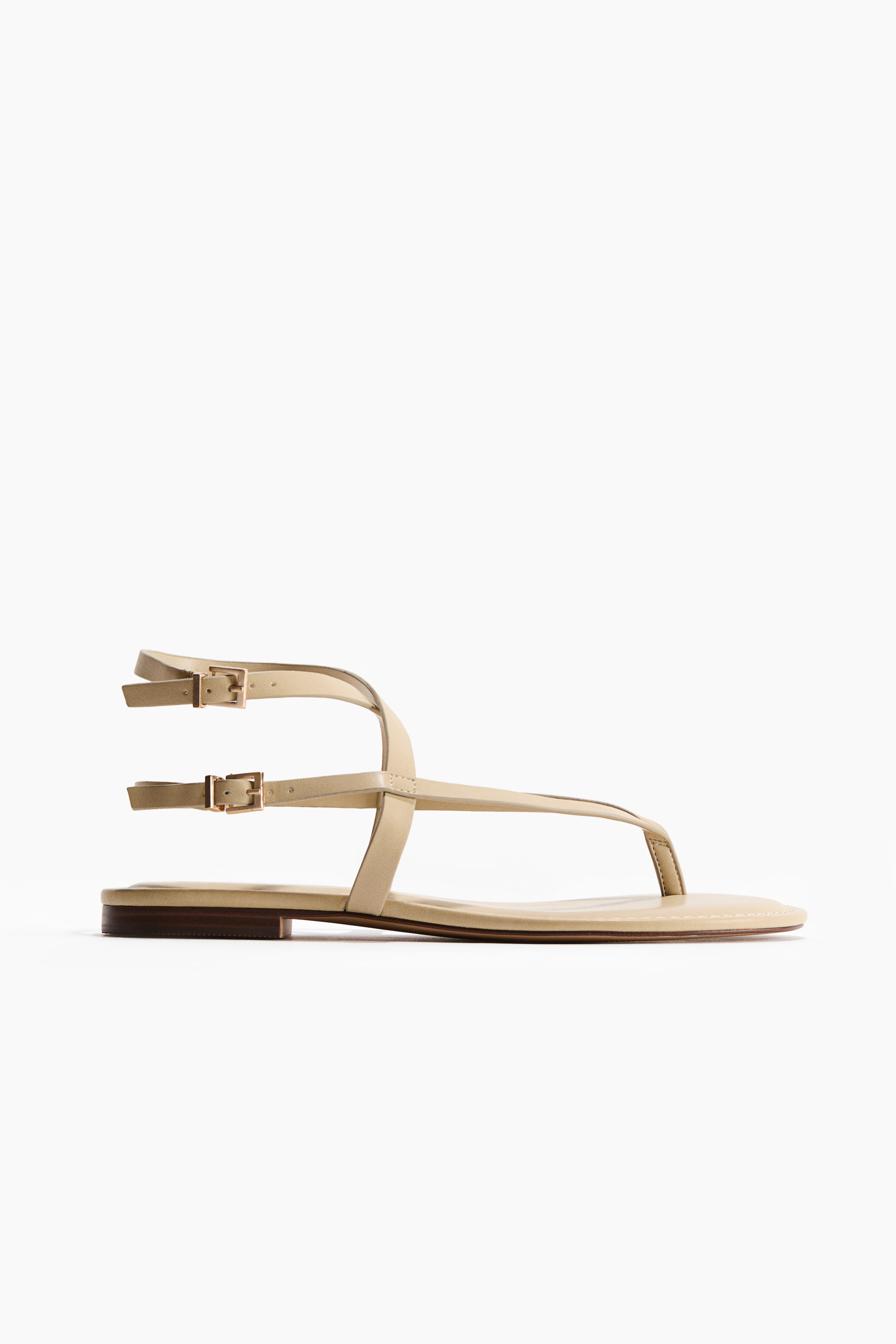 View larger image: A beige flat thong sandal is depicted from a side perspective, showcasing slender straps. One strap runs between the toes, while two delicate straps encircle the ankle, each secured with a gold-toned rectangular buckle. The sandal rests on a flat, thin sole.