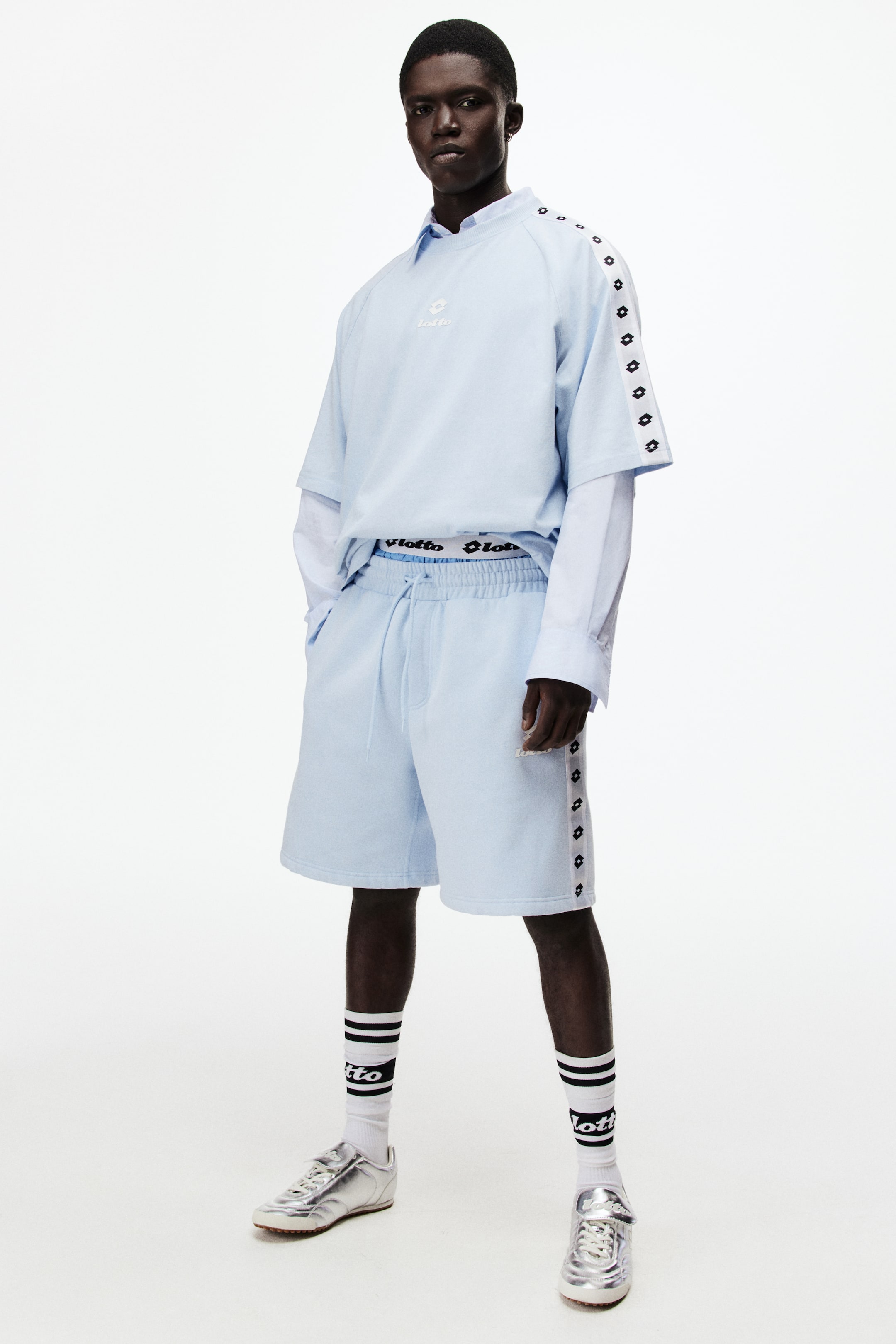 View larger image: The light blue two-piece set, viewed from a front-facing angle, includes a short-sleeved t-shirt and coordinating shorts. The t-shirt showcases a white Lotto logo on the chest and white tape with black diamond logos along the shoulders and sleeves, layered over a light blue collared long-sleeved shirt. The shorts feature an elasticated drawstring waist and white tape with black diamond logos down the sides, with a white waistband displaying "Lotto" branding visible above them.