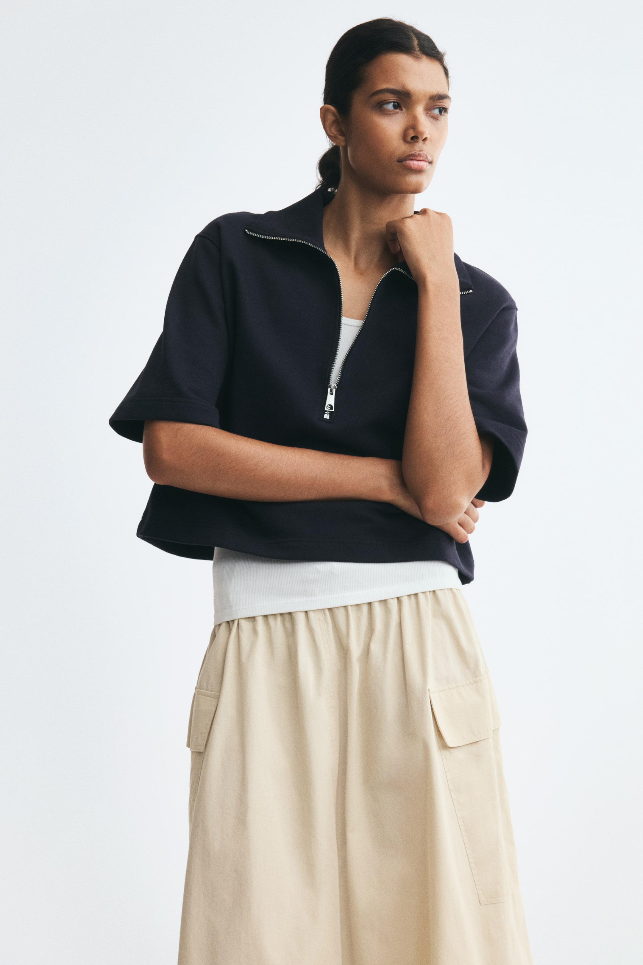 View larger image: A dark blue polo shirt in lightweight sweatshirt fabric, featuring a ribbed collar, half-zip front, dropped shoulders, and elbow-length sleeves, is worn by the model. The cropped-style shirt is layered over a white top and styled with beige trousers.