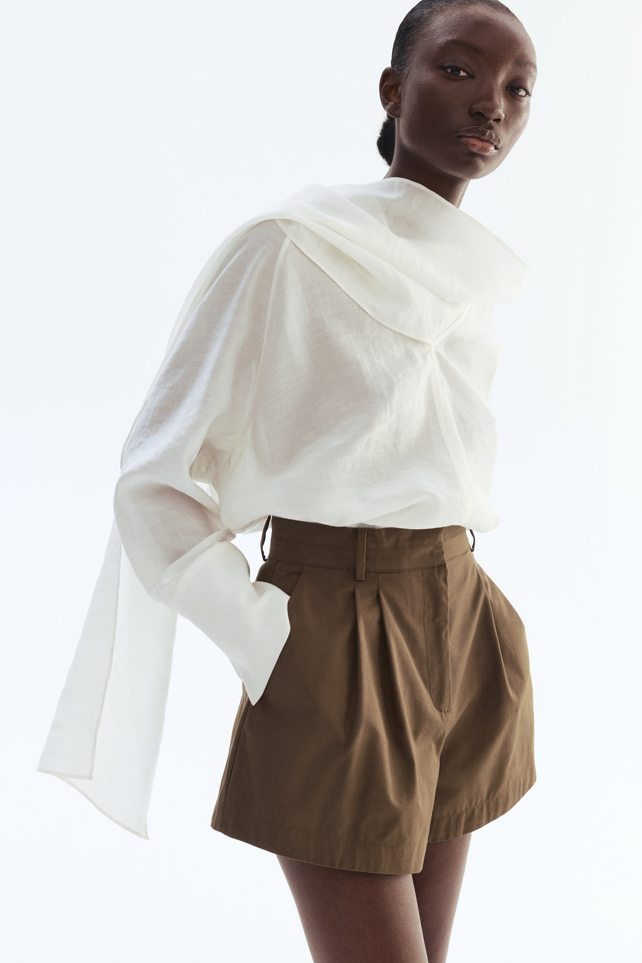 View larger image: The model is showcasing loose-fit, tailored shorts crafted from olive cotton twill. These shorts boast a high waist with front pleats and diagonal side pockets, with the model's left hand casually placed in one of the pockets.