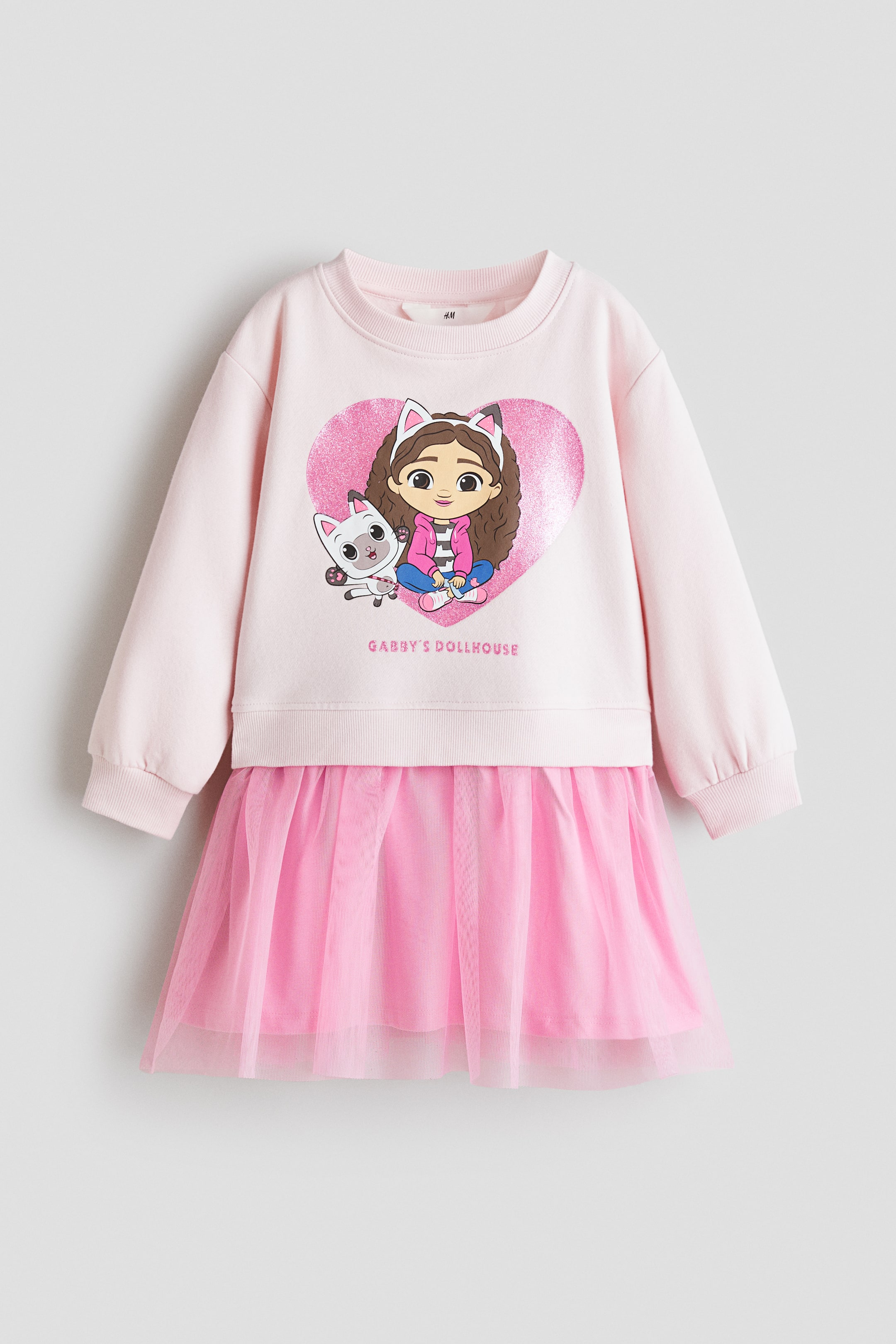 Voir une image plus grande: Une robe rose pâle à manches longues présente un col rond et des poignets côtelés, avec un grand motif de cœur rose scintillant sur le devant du buste montrant une fille de dessin animé et un chat blanc, ainsi que le texte « GABBY'S DOLLHOUSE » en dessous. La jupe est de conception superposée, composée d’une sous-couche rose unie et d’une couche extérieure en tulle rose transparent.