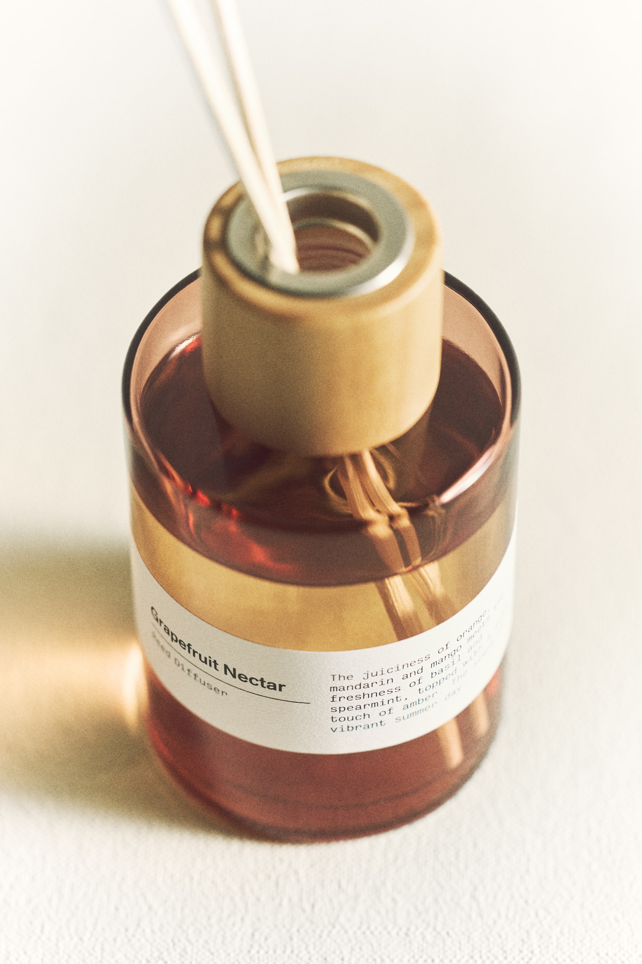 Visa större bild: En doftspridare med en rödbrun cylindrisk glasflaska, sedd från en ovanifrån vinkel. Flaskan är delvis fylld med vätska, och en vit etikett med texten "Grapefruit Nectar Reed Diffuser" sitter på framsidan. En ljusbrun cylindrisk kork med en silverfärgad metallring täcker öppningen, och flera ljusa pinnar sträcker sig uppåt in i vätskan.