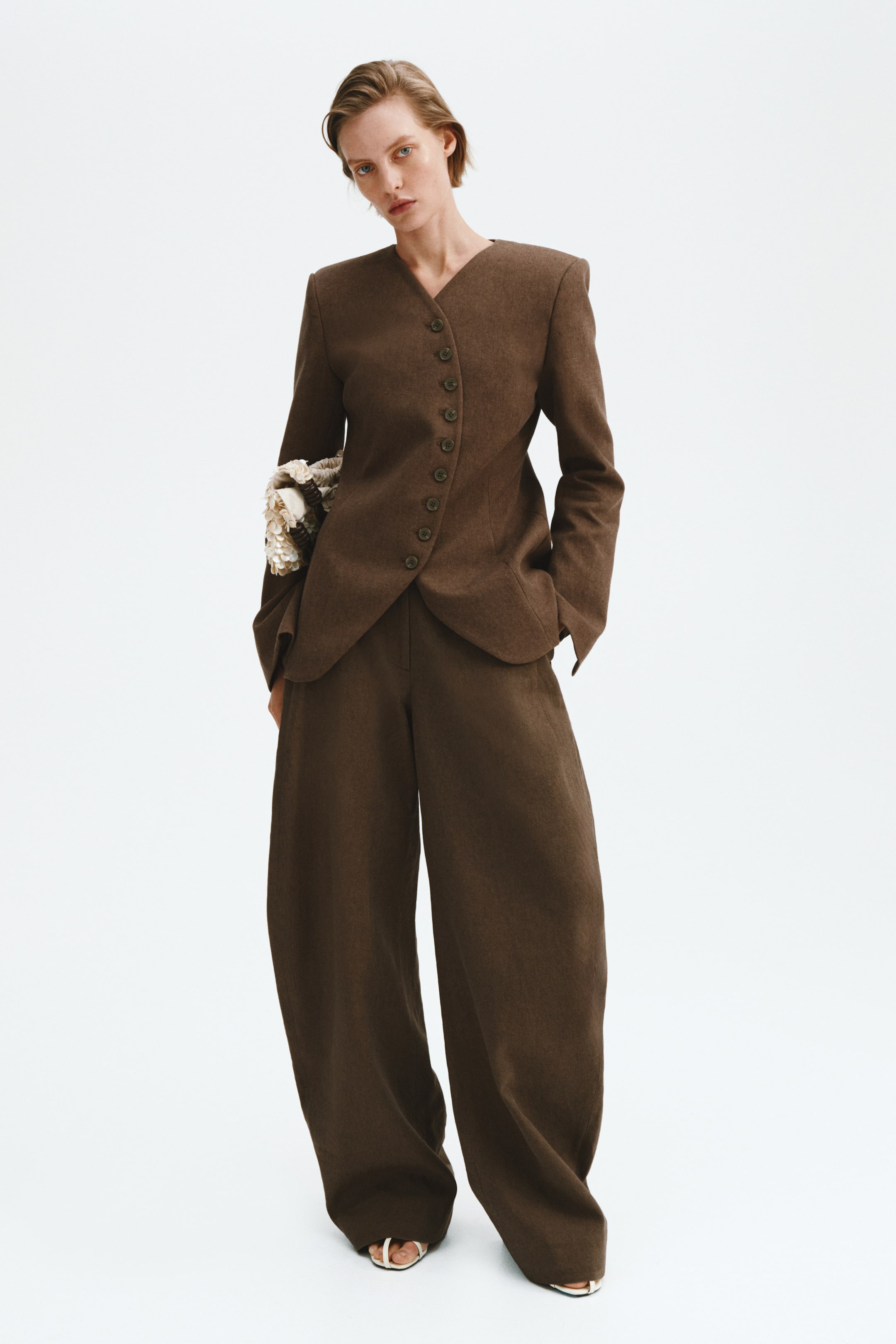 Voir une image plus grande: Un ensemble deux pièces texturé brun comprend un blazer ajusté sans col avec un col en V et une fermeture asymétrique à boutons à l'avant, mettant en valeur des épaules structurées et des manches longues avec des poignets évasés. Le blazer est associé à un pantalon à jambes larges et pleine longueur qui s'étend au-dessus de chaussures blanches à bout ouvert.