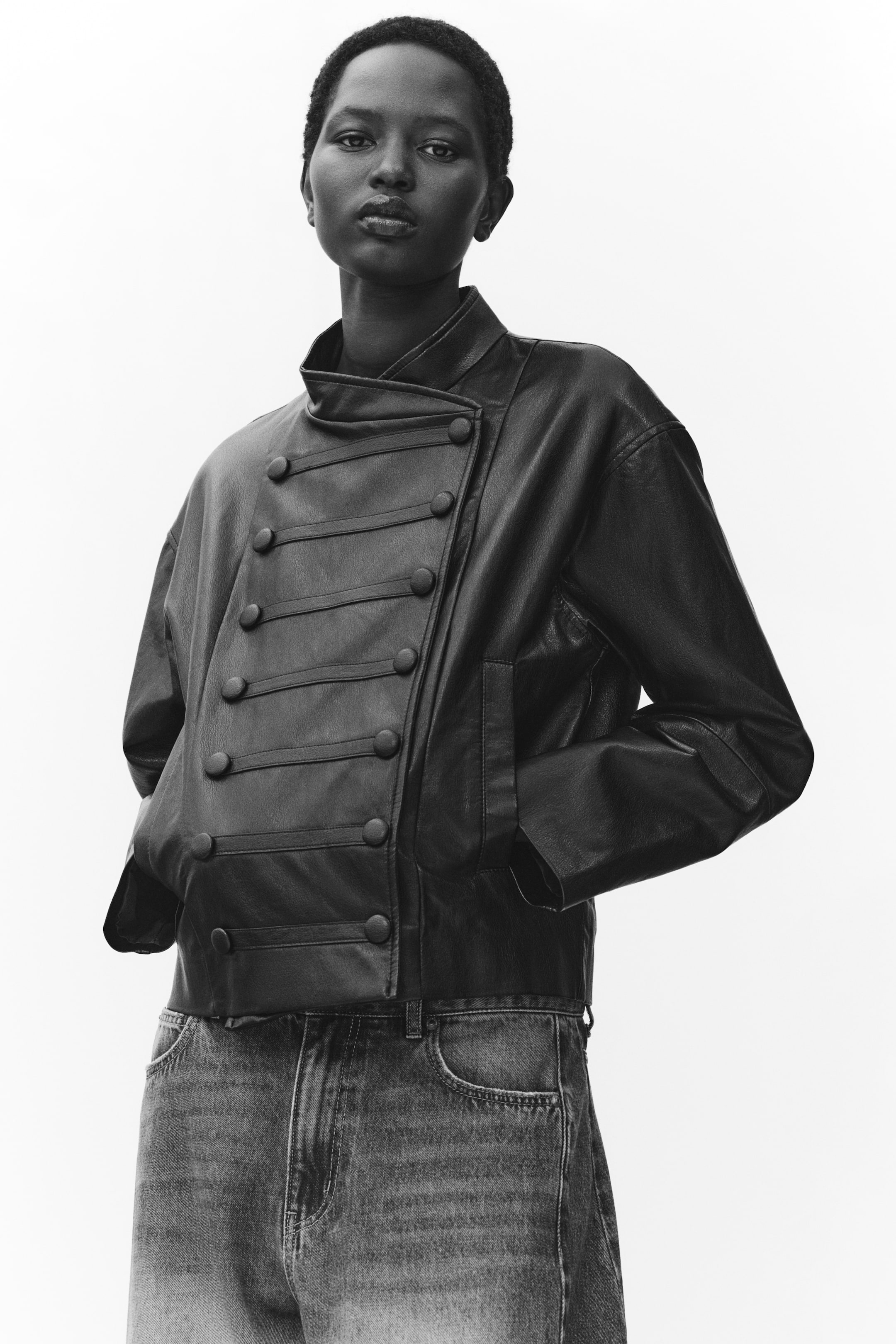 View larger image: The model is wearing a short jacket in coated fabric, with their hands resting inside the welt front pockets. The jacket features a stand collar, an offset front fastening, and decorative horizontal tabs with covered buttons across the front.