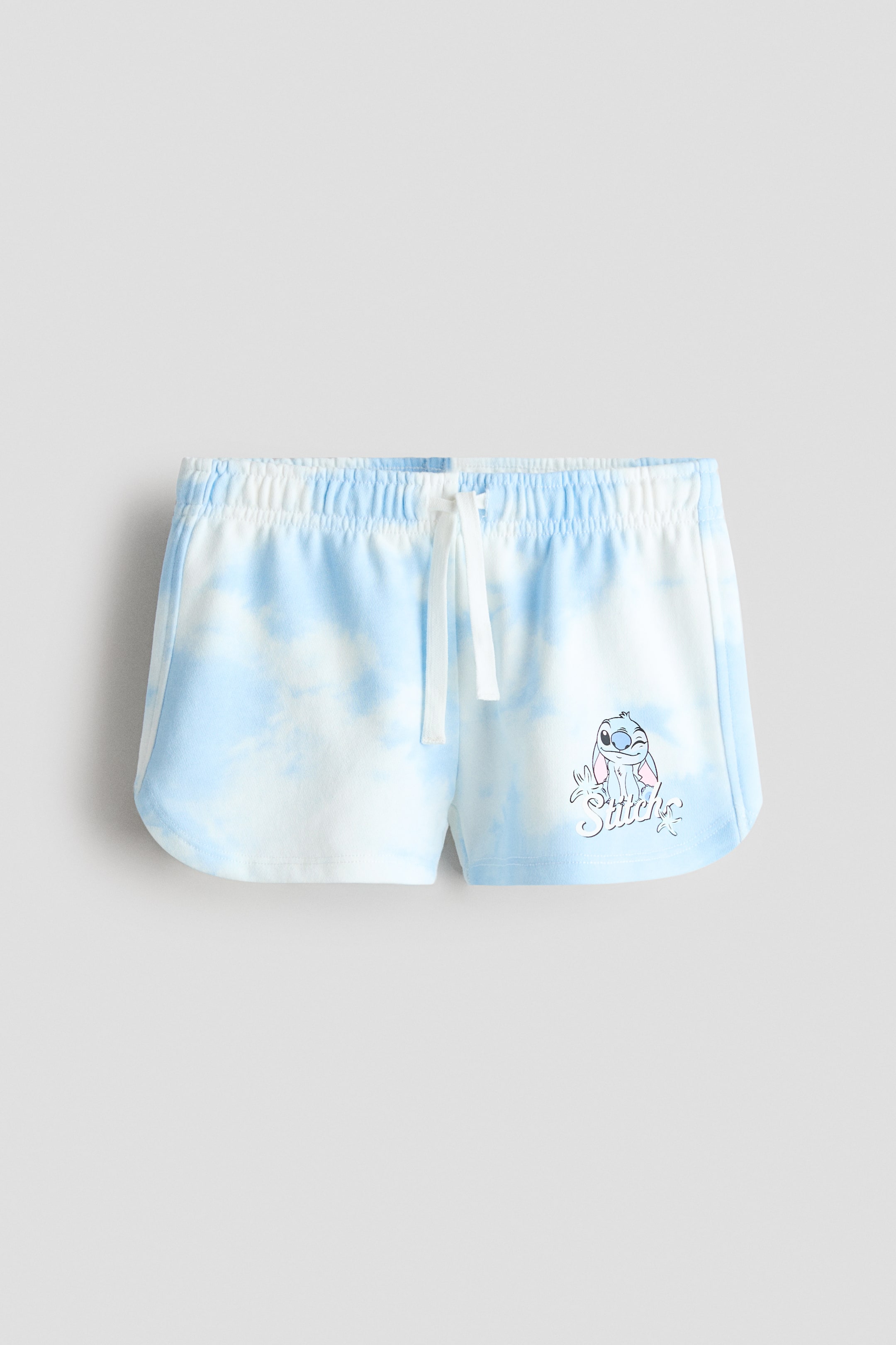 View larger image: The blue and white tie-dye shorts feature an elasticated waistband with a white drawstring tie at the centre front. A graphic of Stitch with the word 'Stitch' appears on the lower left leg, and the leg openings showcase a rounded hem.