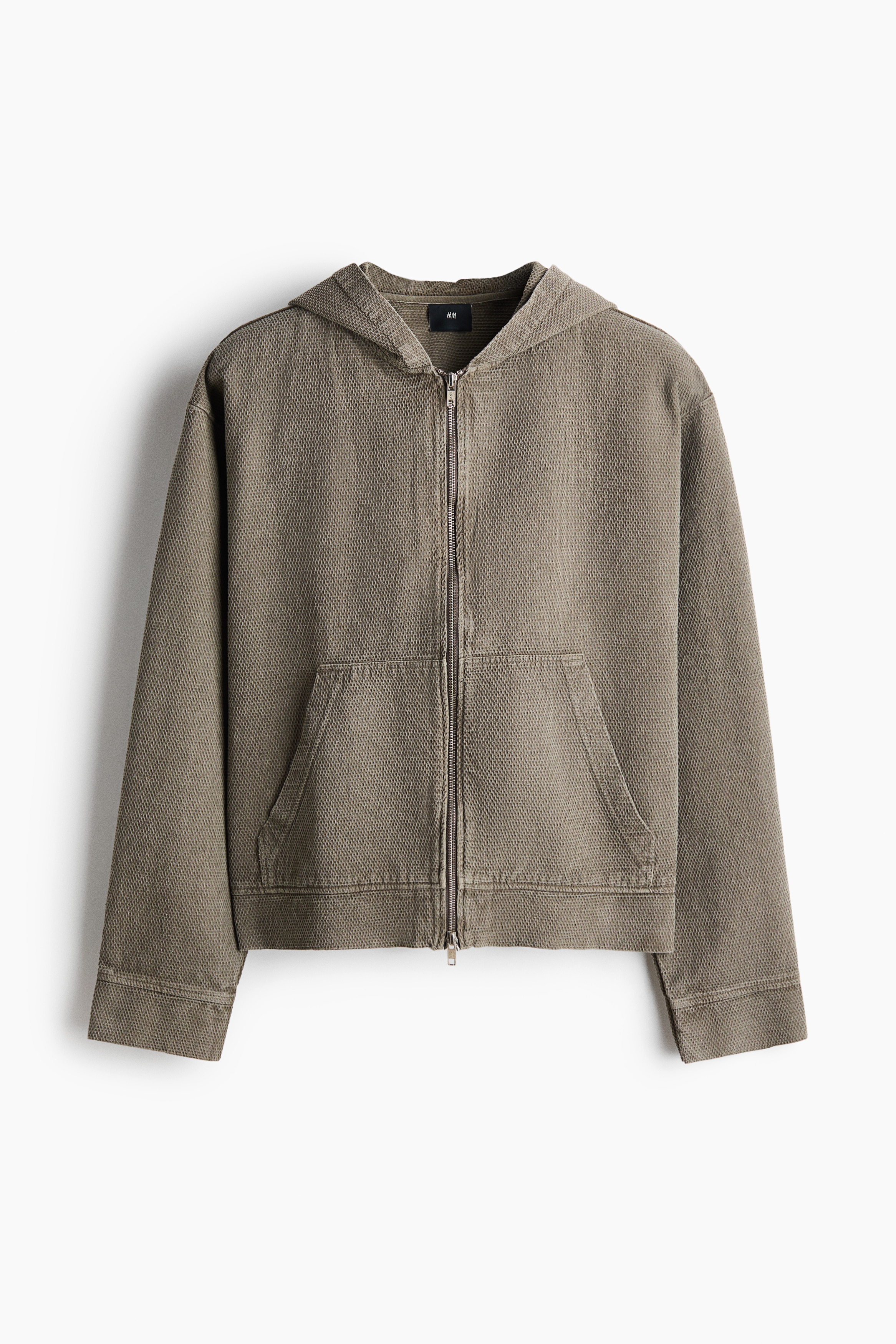 Une surchemise en coton tissÃ© texturÃ©, de couleur brun olive, est portÃ©e par le mannequin, superposÃ©e Ã  un T-shirt blanc et associÃ©e Ã  un pantalon beige. La veste prÃ©sente une capuche non doublÃ©e, une fermeture zippÃ©e frontale Ã  double sens, des Ã©paules tombantes et des manches longues avec poignets.