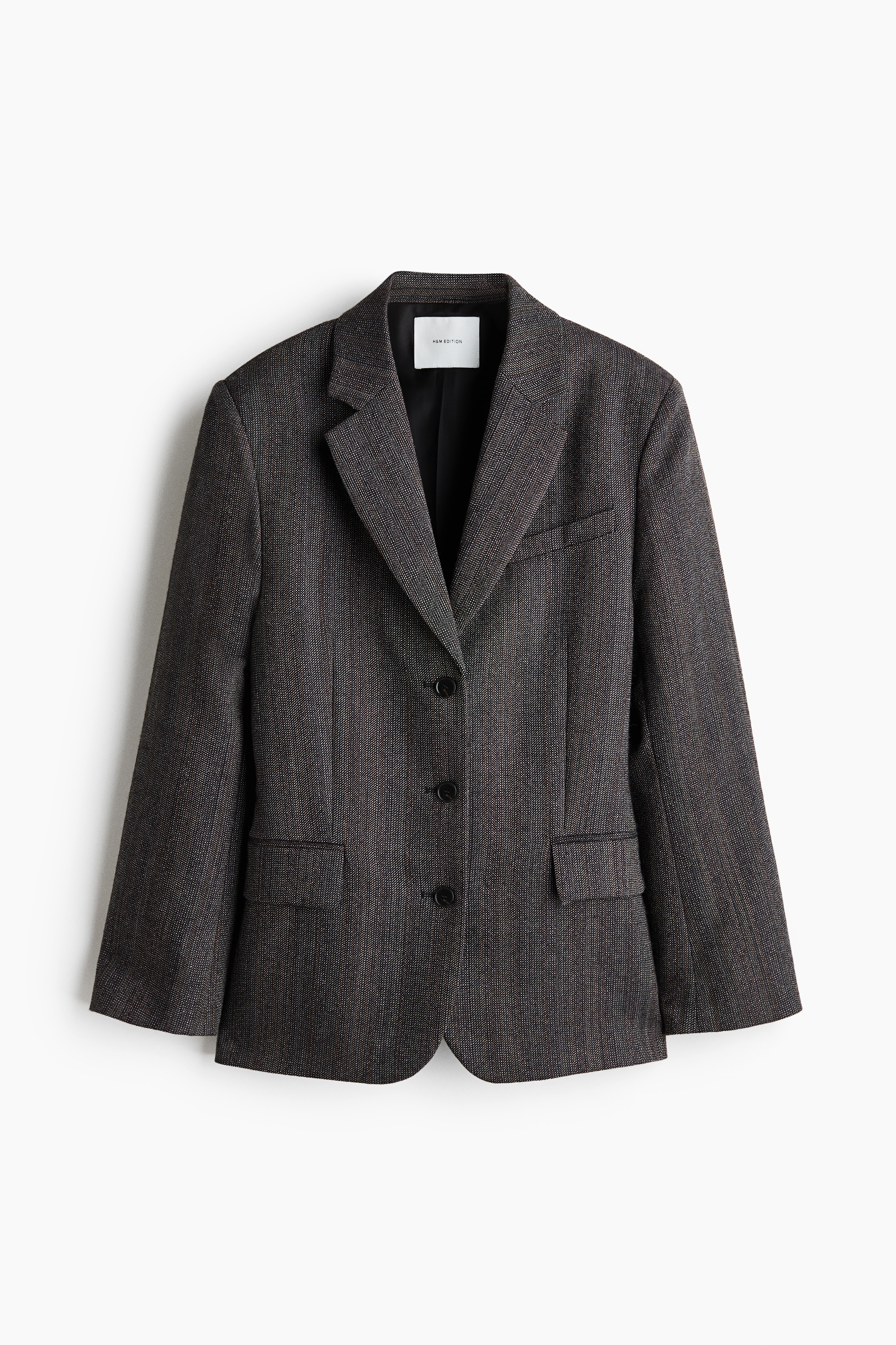 The model is wearing a single-breasted blazer in a textured grey wool weave, layered over a ruched cream blouse. The blazer features structured shoulders, notch lapels, front button fastening, and jetted flap pockets, with the model carrying a small black clutch bag.