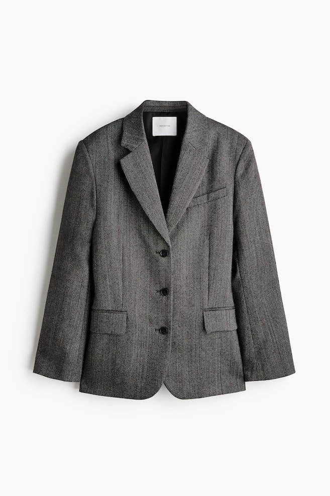 Ein dunkelgrauer, strukturierter Blazer ist von vorne zu sehen und prÃ¤sentiert Revers mit fallender Breite, einen Dreiknopf-Verschluss vorne und ausgeprÃ¤gte Schultern. Der Blazer zeigt zwei untere Pattentaschen und eine einzelne Brustleistentasche. - 1