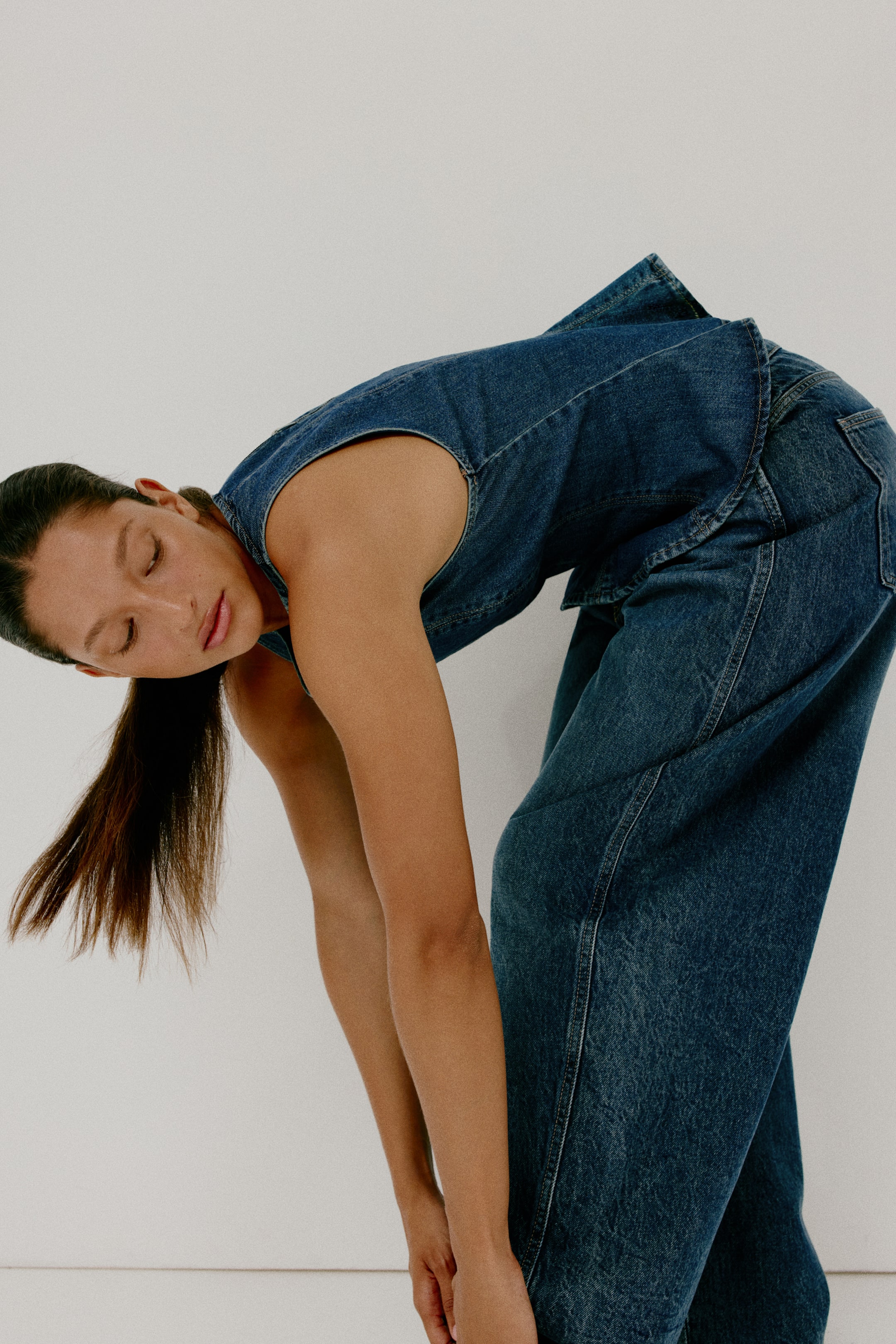 View larger image: A dark wash denim sleeveless jumpsuit is being showcased from a side-back angle, featuring a relaxed fit. The wide-leg trousers display prominent side seams, and a folded fabric detail is visible at the upper hip.