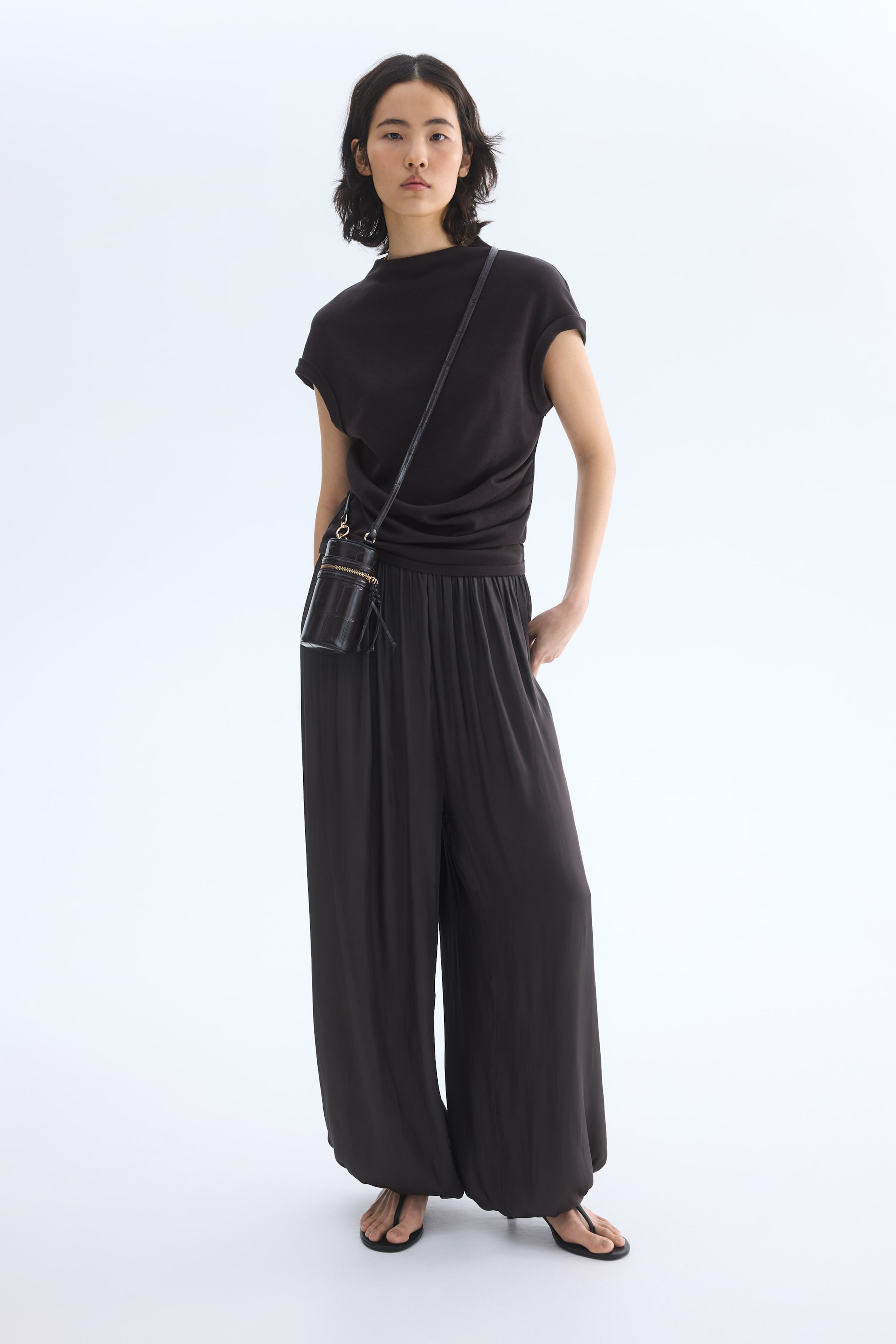 Προβολή μεγαλύτερης εικόνας: Μια μαύρη κυλινδρική τσάντα crossbody με ανάγλυφη υφή κροκόδειλου και φερμουάρ σε χρυσή απόχρωση, φοριέται διαγώνια στο σώμα.