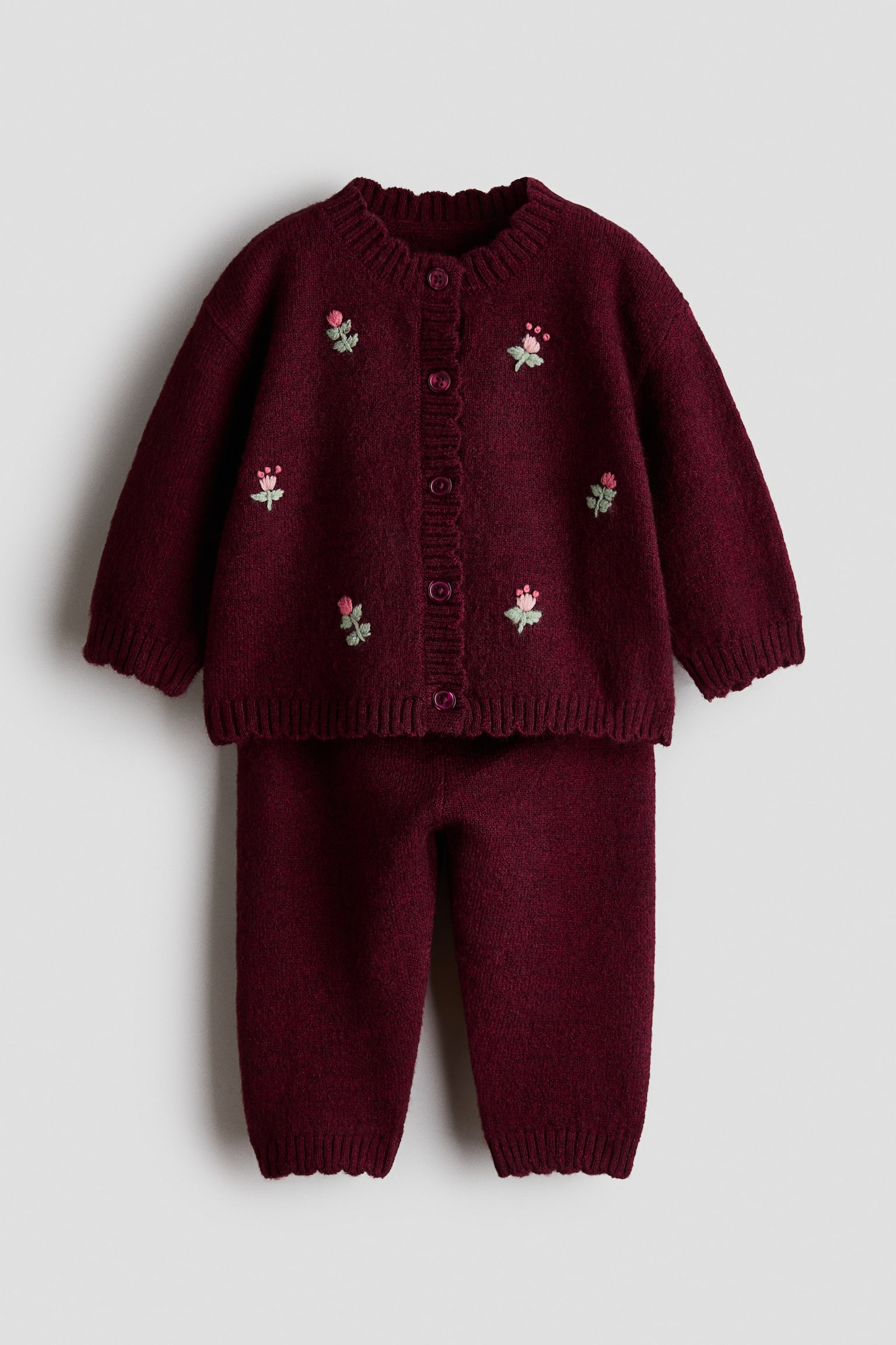 Un ensemble deux piÃ¨ces en tricot bordeaux est prÃ©sentÃ© de face, comprenant un cardigan boutonnÃ© ornÃ© de petites fleurs roses brodÃ©es et un pantalon en tricot assorti. Le cardigan affiche un col rond cÃ´telÃ© ainsi que des poignets et un ourlet cÃ´telÃ©s, tandis que le pantalon prÃ©sente des chevilles cÃ´telÃ©es.