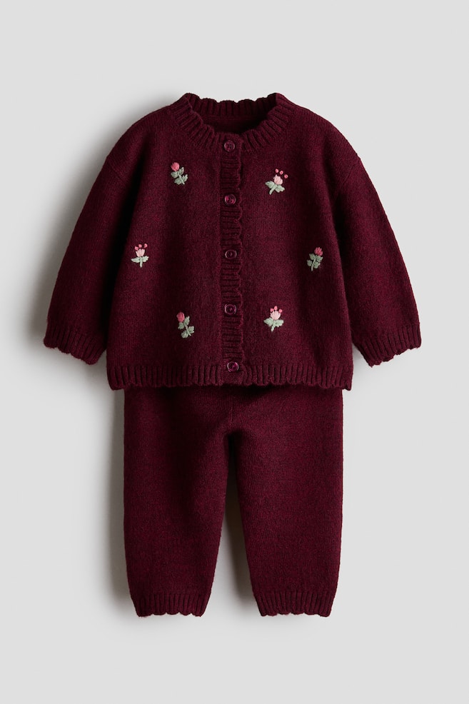 A burgundy knitted two-piece set is shown from the front, featuring a button-down cardigan with small embroidered pink flowers and matching knitted trousers. The cardigan features a ribbed crew neckline, cuffs, and hem, while the trousers have ribbed ankle cuffs. - 1