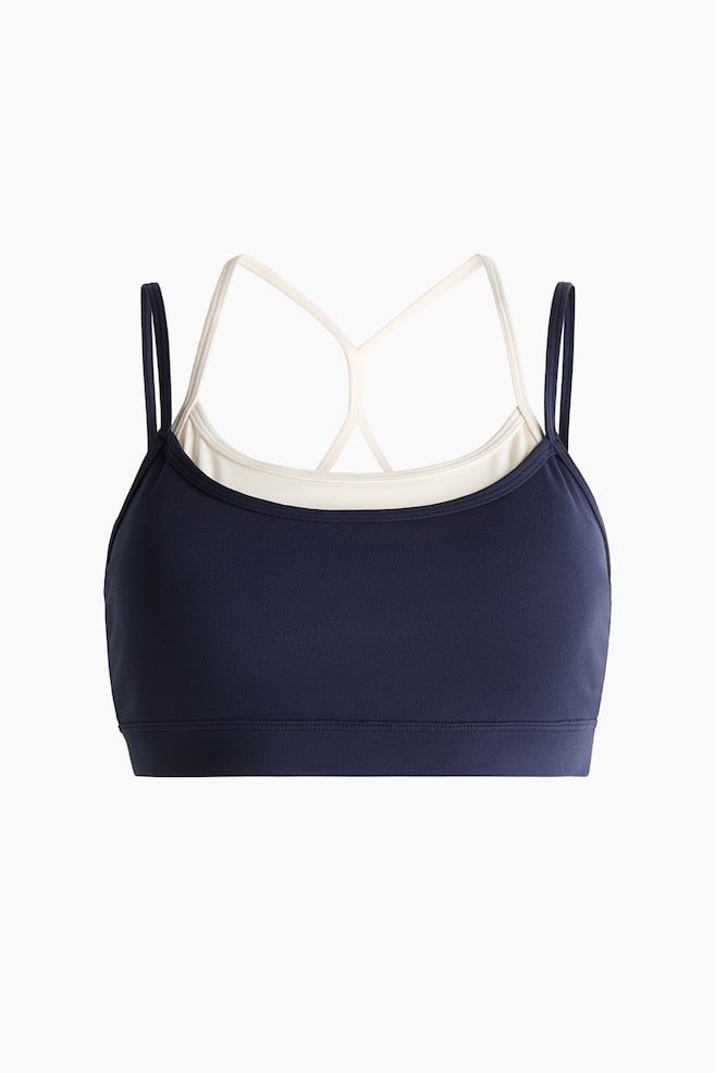 "Sports bra with SoftMove? showcases a sleek front design. The navy blue outer layer features delicate spaghetti straps, overlaying a light-coloured inner layer with thin cross-back straps. A wide stitched bottom band completes the look." - 1