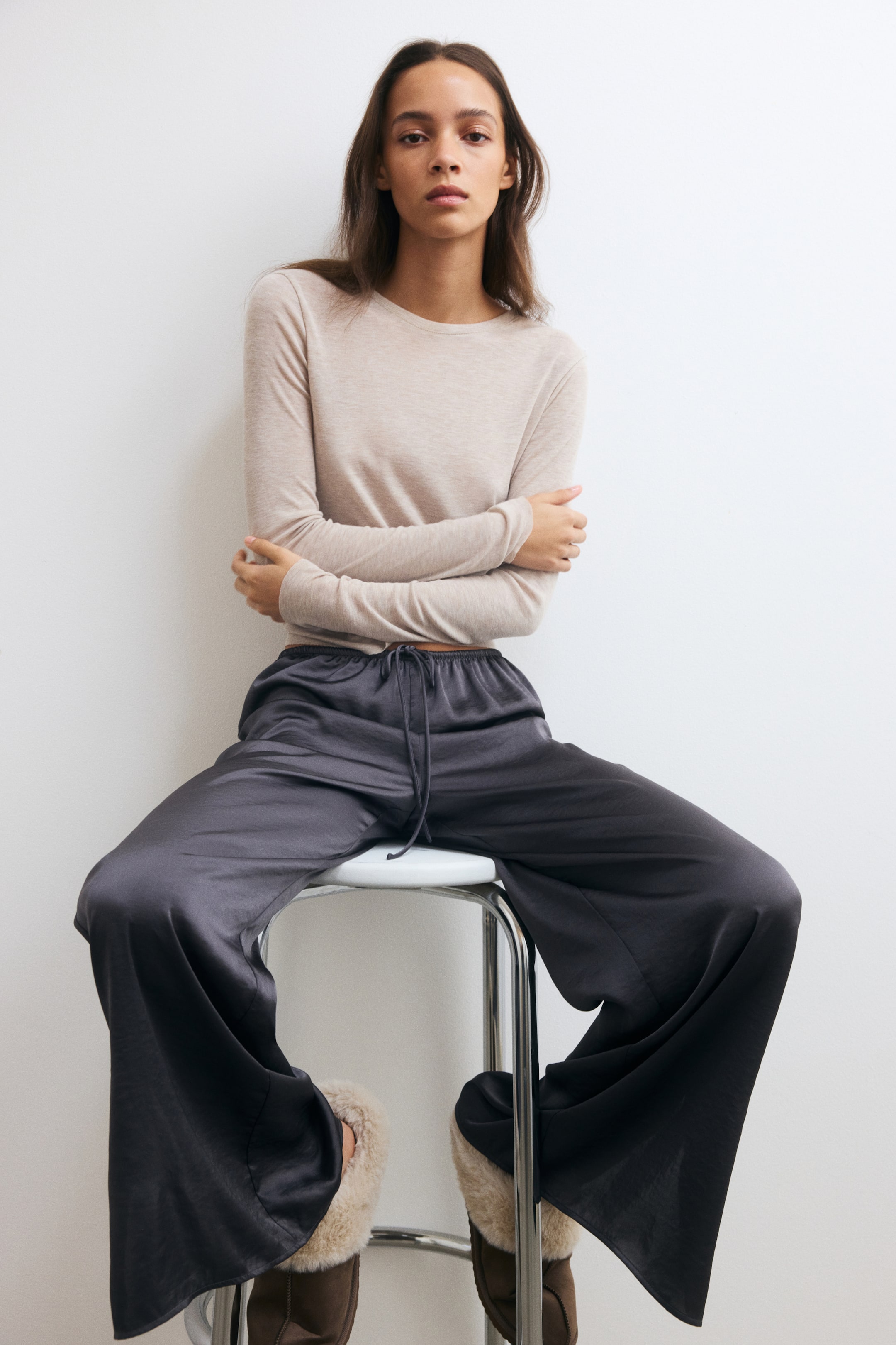 Grösseres Bild ansehen: Ein Pyjama-Set mit einem hellbeigen, langärmligen Ripp-Jersey-Top mit Rundhalsausschnitt und dunkelgrauen, weit geschnittenen Satin-Hosen mit elastischem Bund und Kordelzug wird vom Model getragen. Das Model sitzt mit verschränkten Armen auf einem Hocker und trägt das Set zusammen mit braunen, mit Plüsch gefütterten Hausschuhen.
