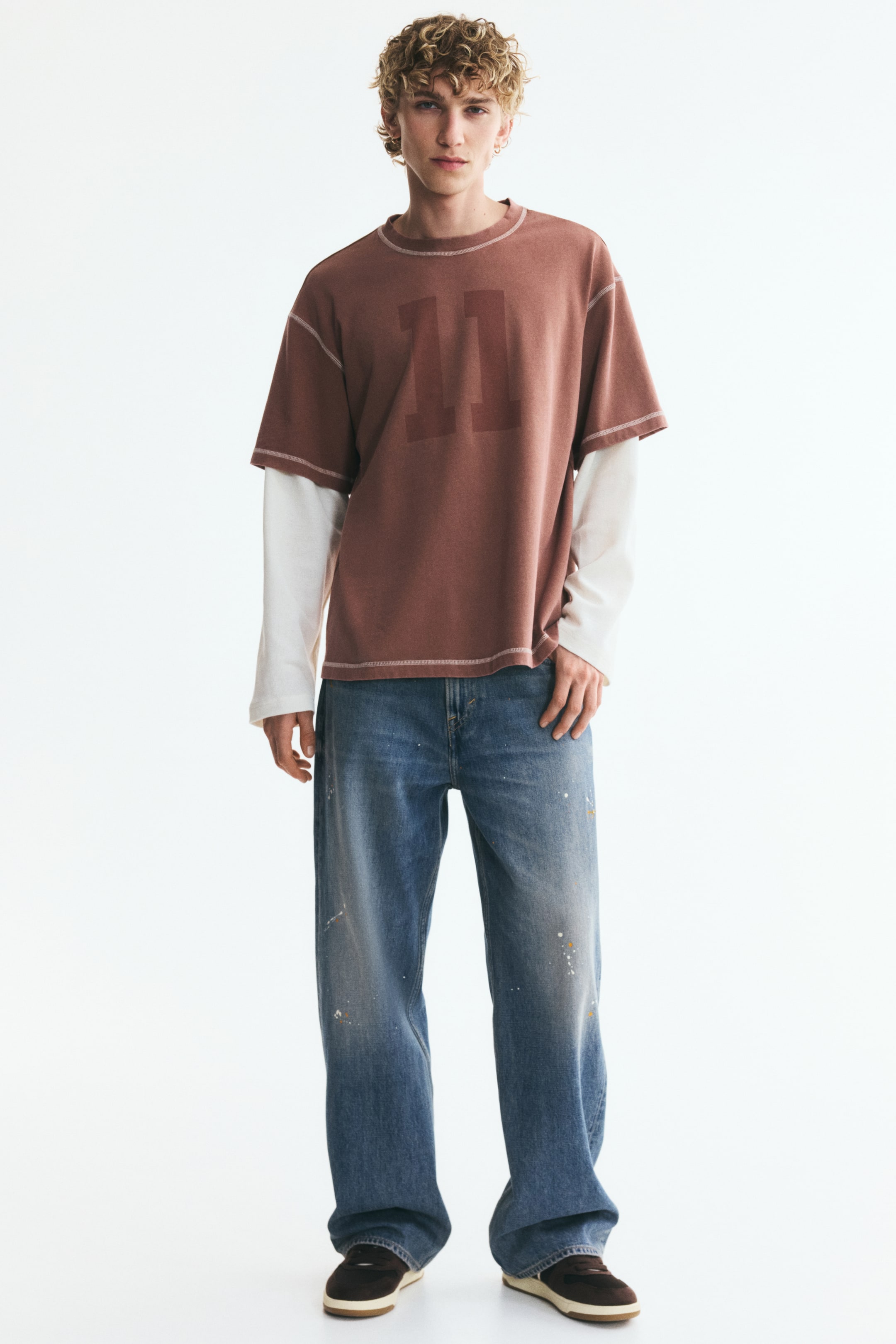 View larger image: Loose-fit, straight-leg blue denim jeans featuring a dropped crotch and subtle paint splatters are worn by the model. The jeans are paired with a layered brown and white long-sleeve T-shirt and dark sneakers.