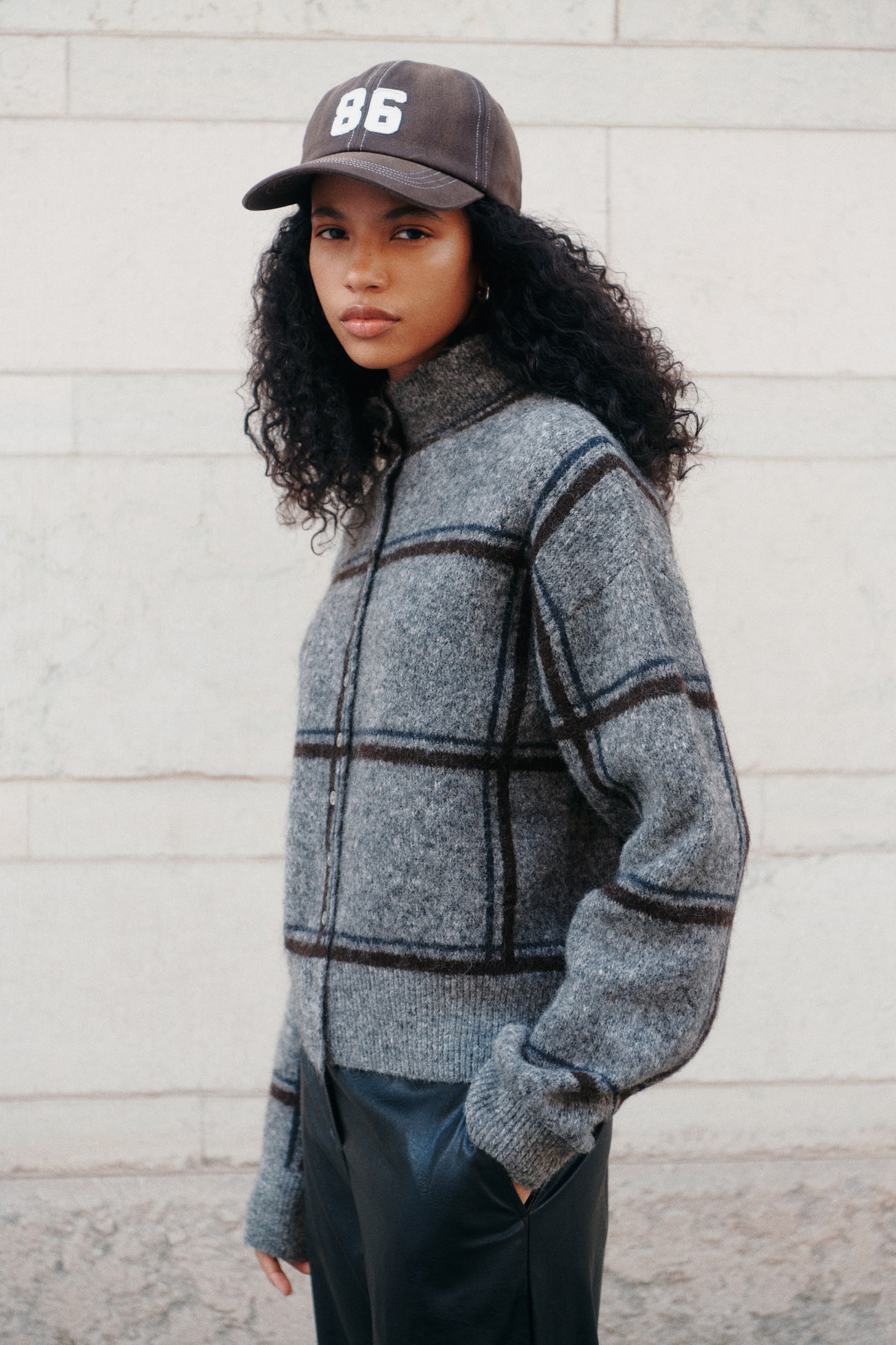 Agrandir l'image: Un cardigan en maille à motif écossais gris, doté d’un col montant et d’une fermeture boutonnée sur le devant, est porté par le mannequin. Le cardigan présente des poignets et un ourlet côtelés, et est assorti à un pantalon foncé ainsi qu’à une casquette de baseball marron.
