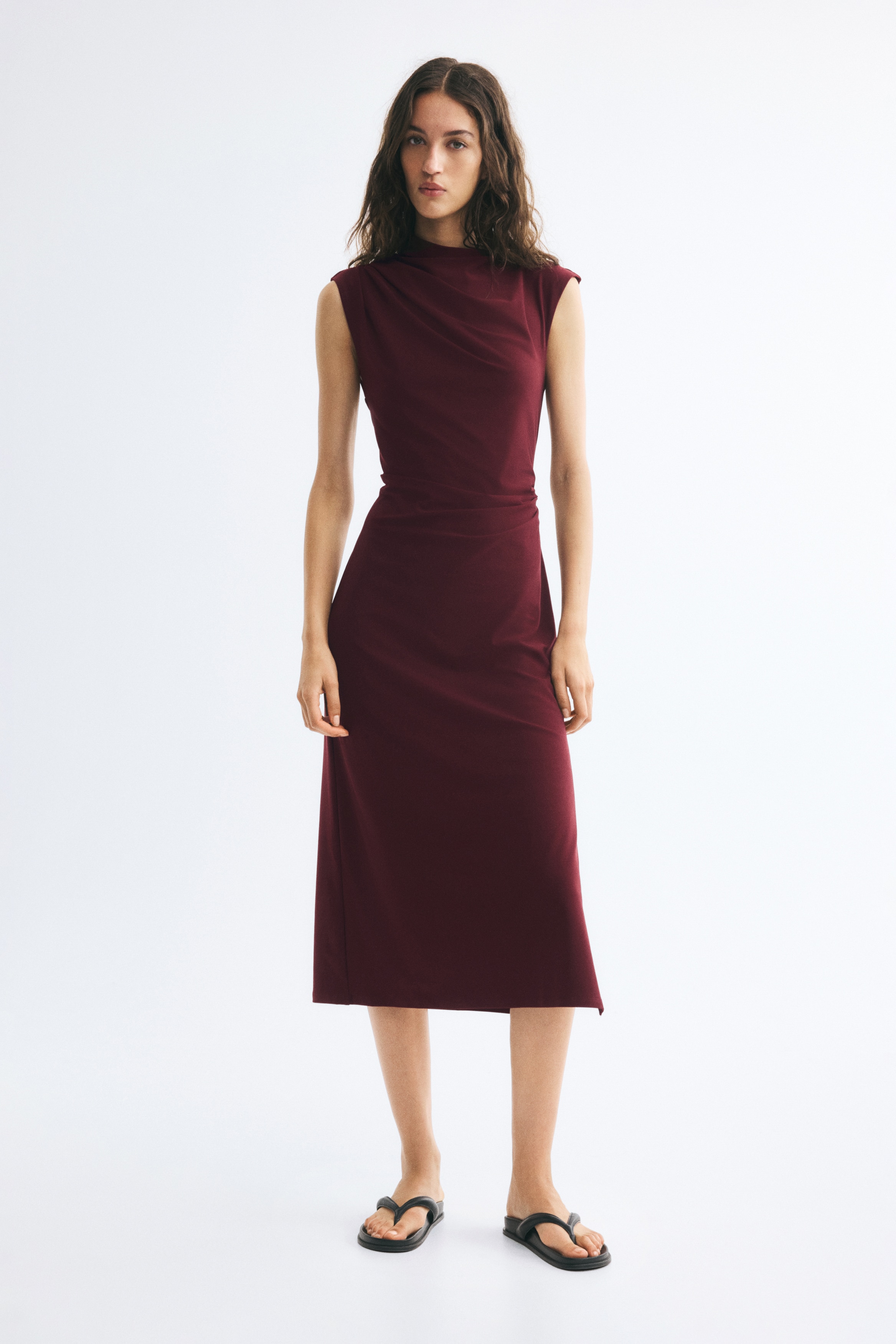 Má»t chiáº¿c vÃ¡y midi khÃ´ng tay mÃ u Äá» burgundy Äang ÄÆ°á»£c treo hÆ°á»ng máº·t trÆ°á»c, ná»i báº­t vá»i cá» Ã¡o xáº¿p náº¿p báº¥t Äá»i xá»©ng vÃ  chi tiáº¿t nhÃºn táº¡i eo, táº¡o nÃªn nhá»¯ng náº¿p gáº¥p buÃ´ng rá»§ thanh lá»ch dá»c theo chiá»u dÃ i trang phá»¥c.