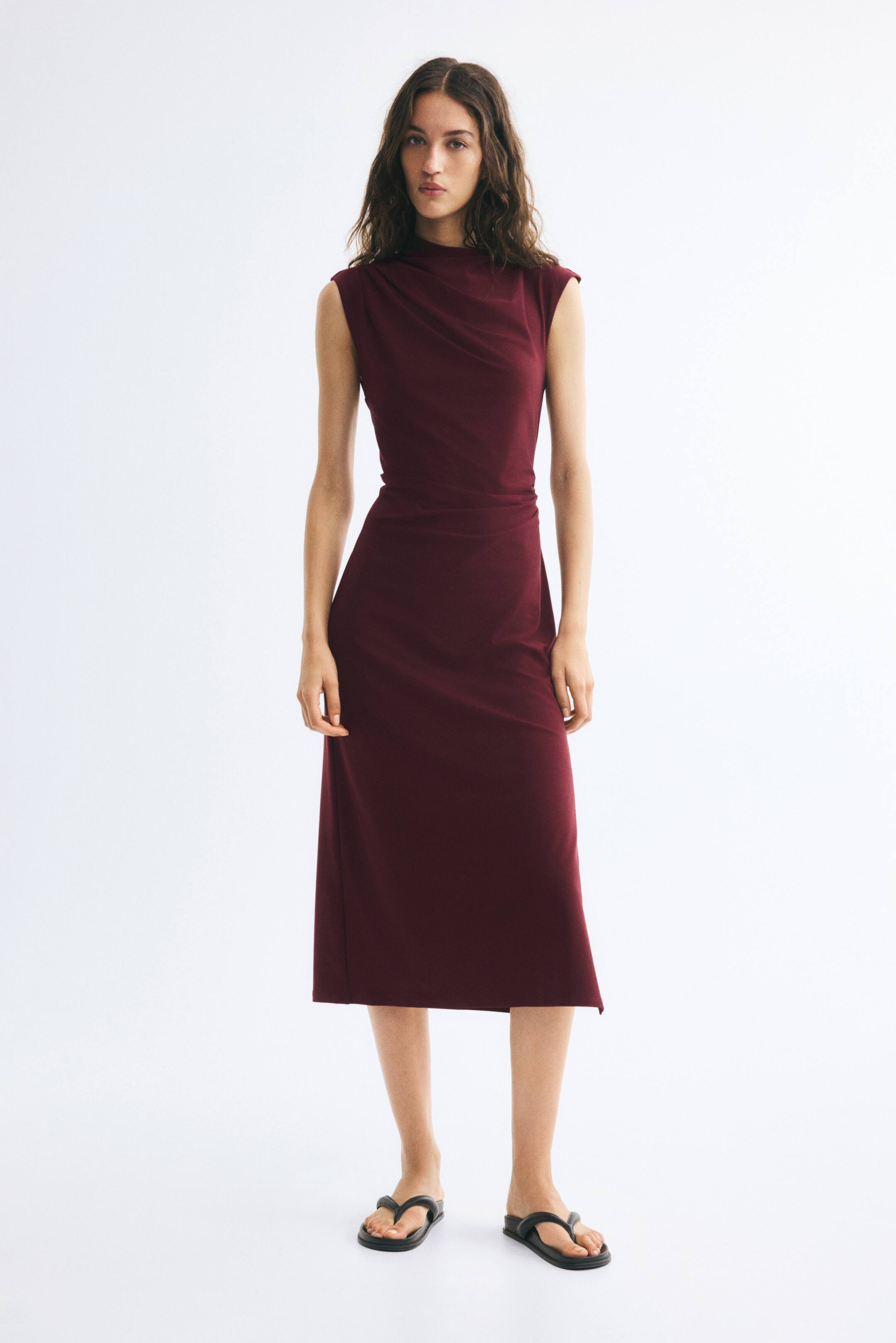 Xem ảnh lớn hơn: Người mẫu đang mặc một chiếc váy jersey màu đỏ burgundy, không tay, dài đến bắp chân, ôm dáng, với phần cổ lệch vai kiểu drape bất đối xứng và chi tiết nhún ở eo. Váy được phối cùng sandals xỏ ngón đế bệt màu đen.
