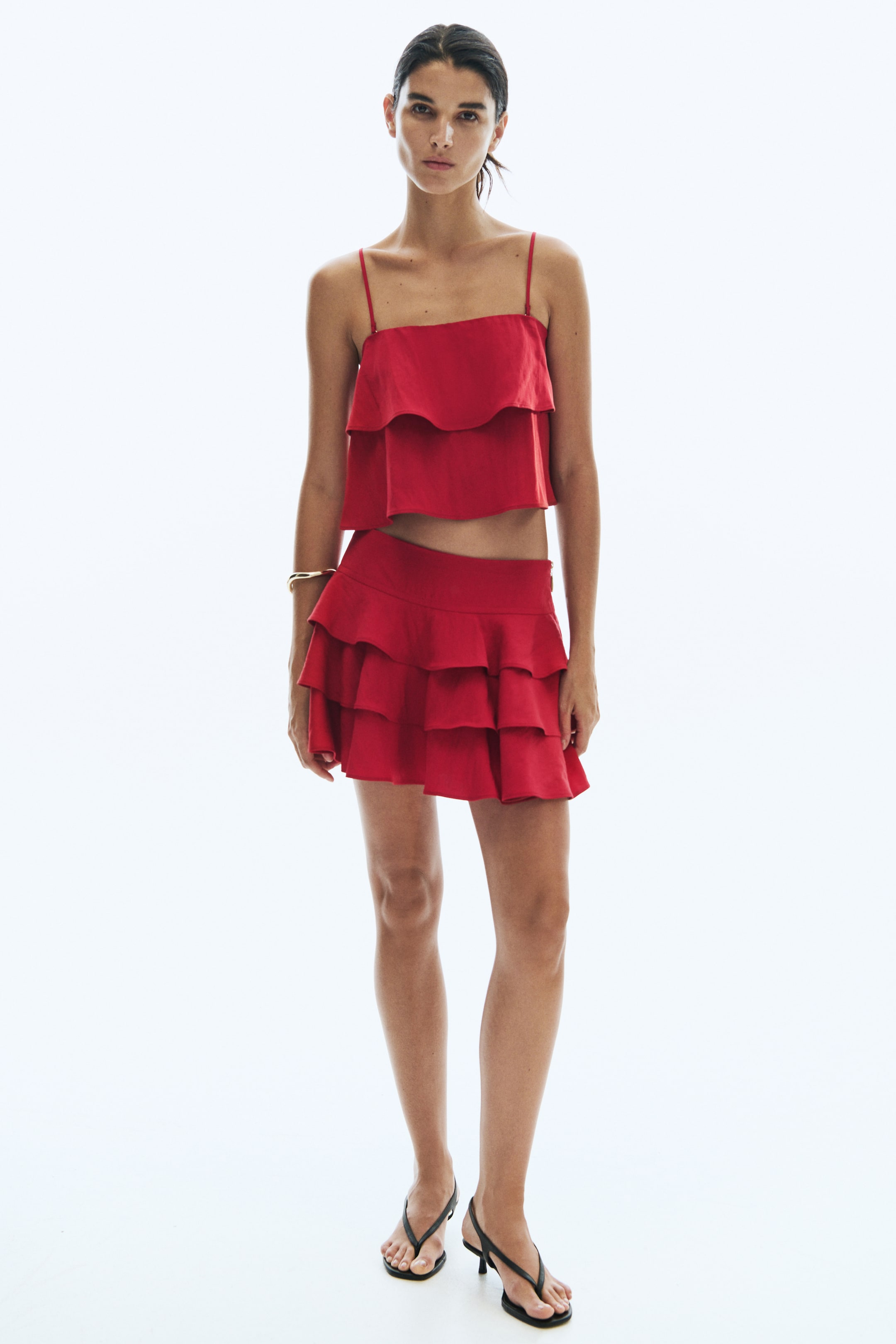 View larger image: A red mini skirt featuring a double-layered waistband and three flounced tiers is being worn by the model. The skirt is paired with a matching red cropped camisole top that also features ruffled layers.