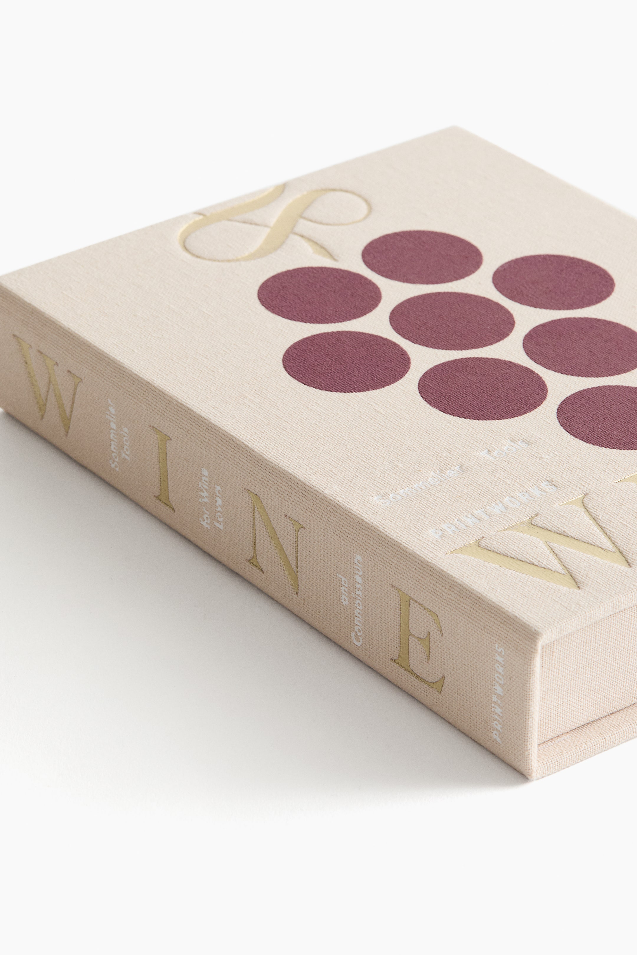 Ver imagem maior: Um livro inclinado com uma capa de tecido bege texturizado está a exibir o seu dorso, mostrando 'WINE' em grandes letras douradas, juntamente com texto dourado menor: 'Sommelier Tools For Wine Lovers and Connoisseurs' e 'PRINTWORKS'. A sua capa frontal apresenta um conjunto de nove círculos cor de vinho e uma parte de um grande gráfico dourado 'S'.