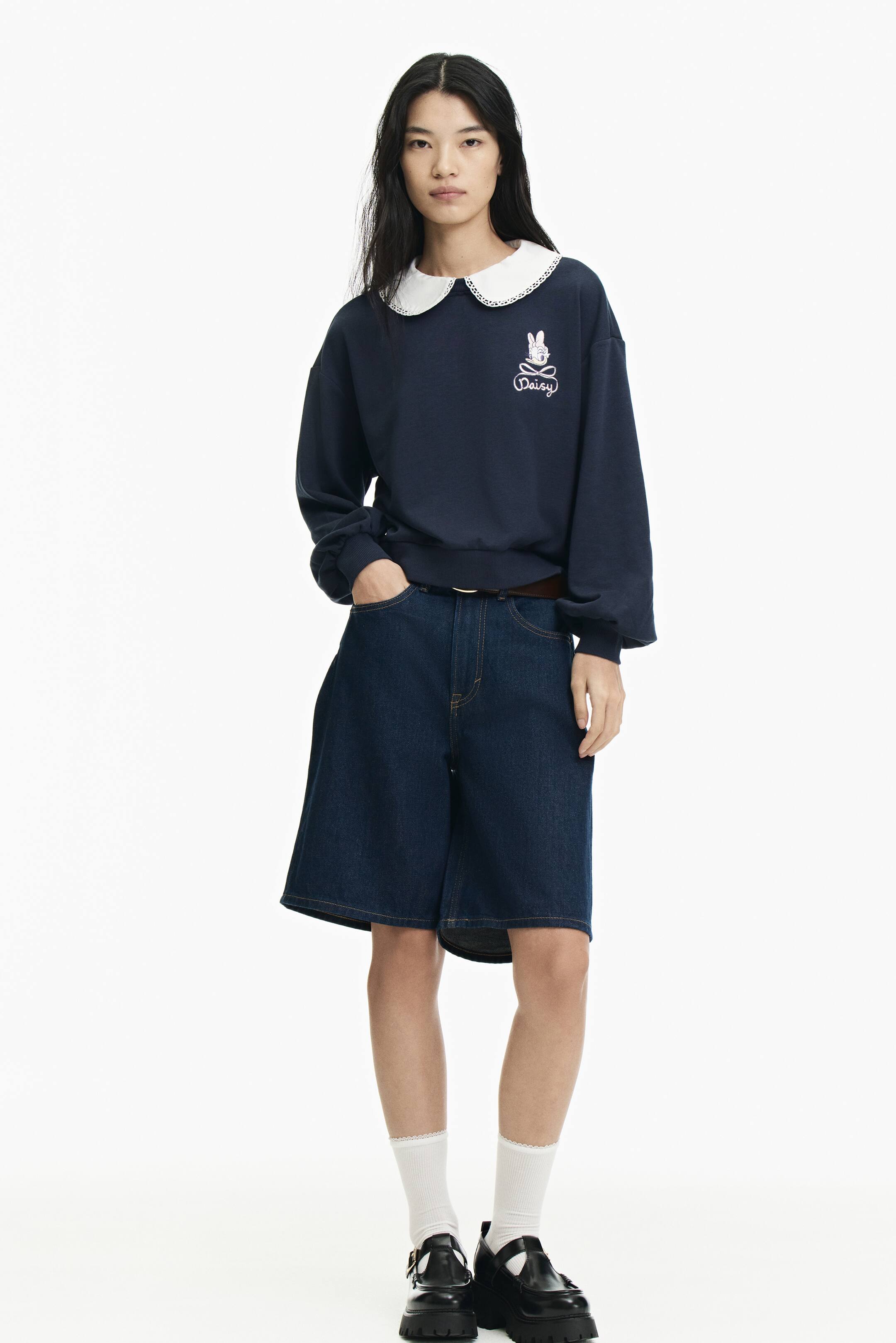 Xem ảnh lớn hơn: Người mẫu đang mặc một áo top màu xanh navy bằng chất liệu sweatshirt mỏng nhẹ. Thiết kế nổi bật với họa tiết thỏ thêu cùng chữ “Daisy” ở ngực, cổ áo cotton lớn viền ren trắng, vai trễ và tay dài phồng với bo tay cùng gấu áo bo rib. Người mẫu đang đứng với một tay đặt trong túi.