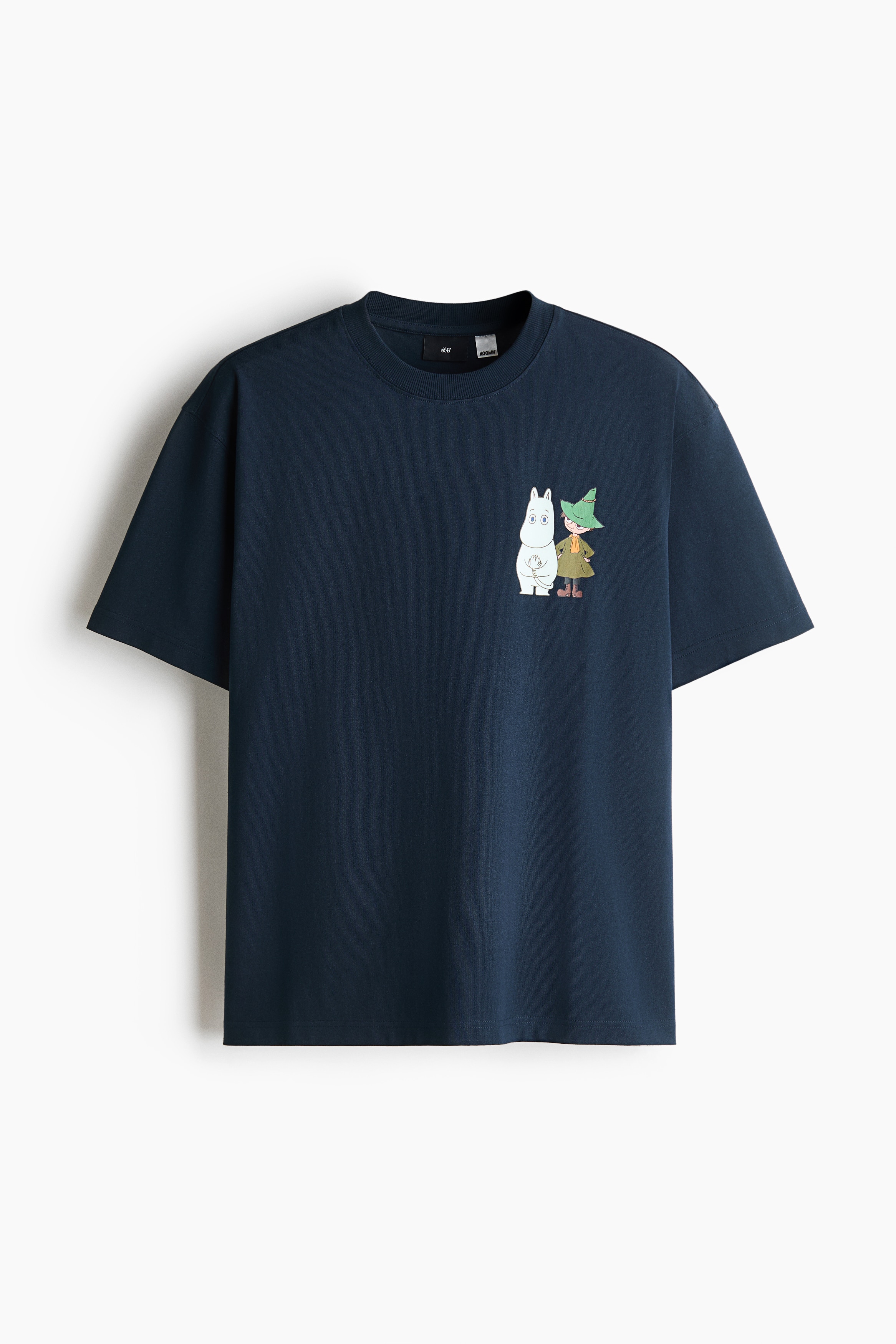 A navy blue, loose-fit T-shirt featuring a small graphic print on the left chest is worn by the model over a layered white T-shirt. The T-shirt has a ribbed crew neck and dropped shoulders, and is styled with blue denim jeans.