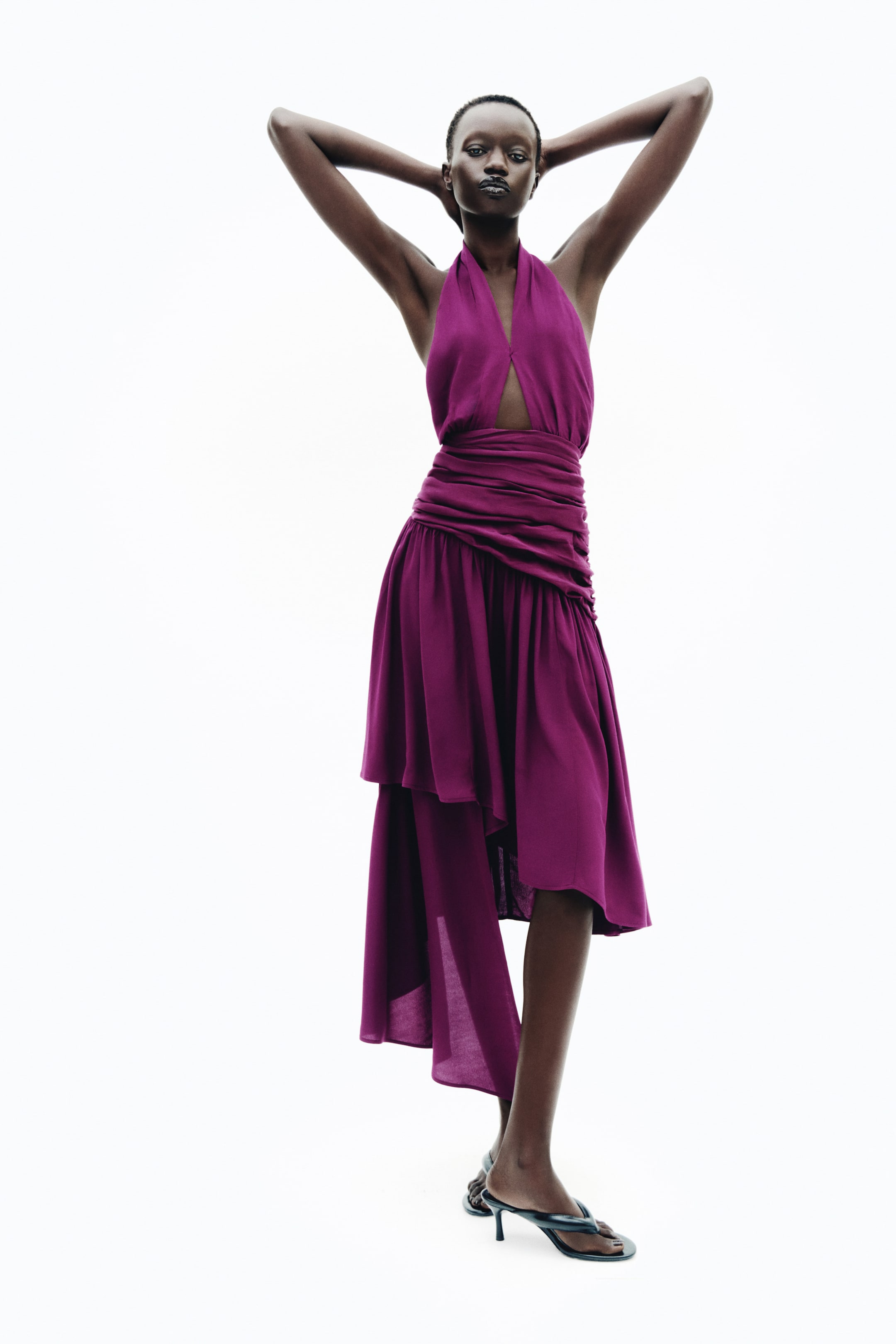 Voir une image plus grande: Une robe licou violette jusqu’au mollet est portée par le mannequin, avec une découpe à l’avant au niveau du buste et un panneau de taille fortement froncé. La robe s’évaser en une jupe fluide à ourlet asymétrique spectaculaire et est agencée avec des sandales noires à talons.