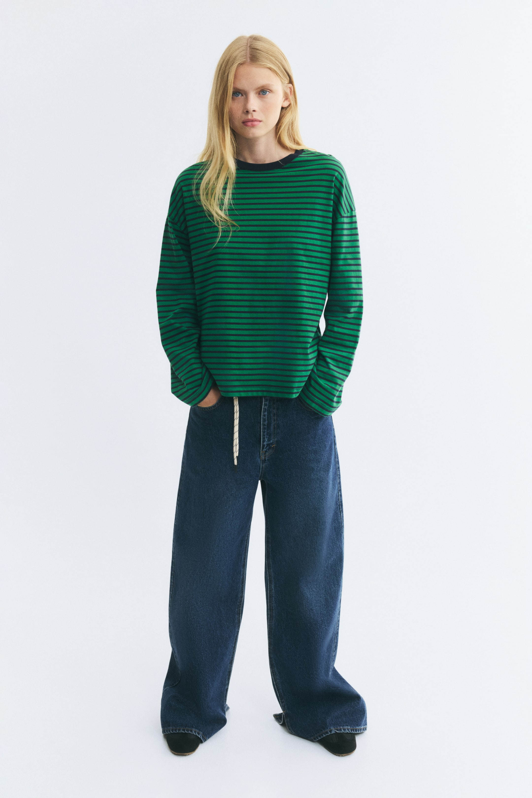 Visualizza l'immagine ingrandita: La modella indossa un top a maniche lunghe a righe orizzontali verdi e nere, con scollo rotondo scuro e spalle scese. La modella è in piedi con le mani infilate nelle tasche anteriori di jeans blu a gamba larga.