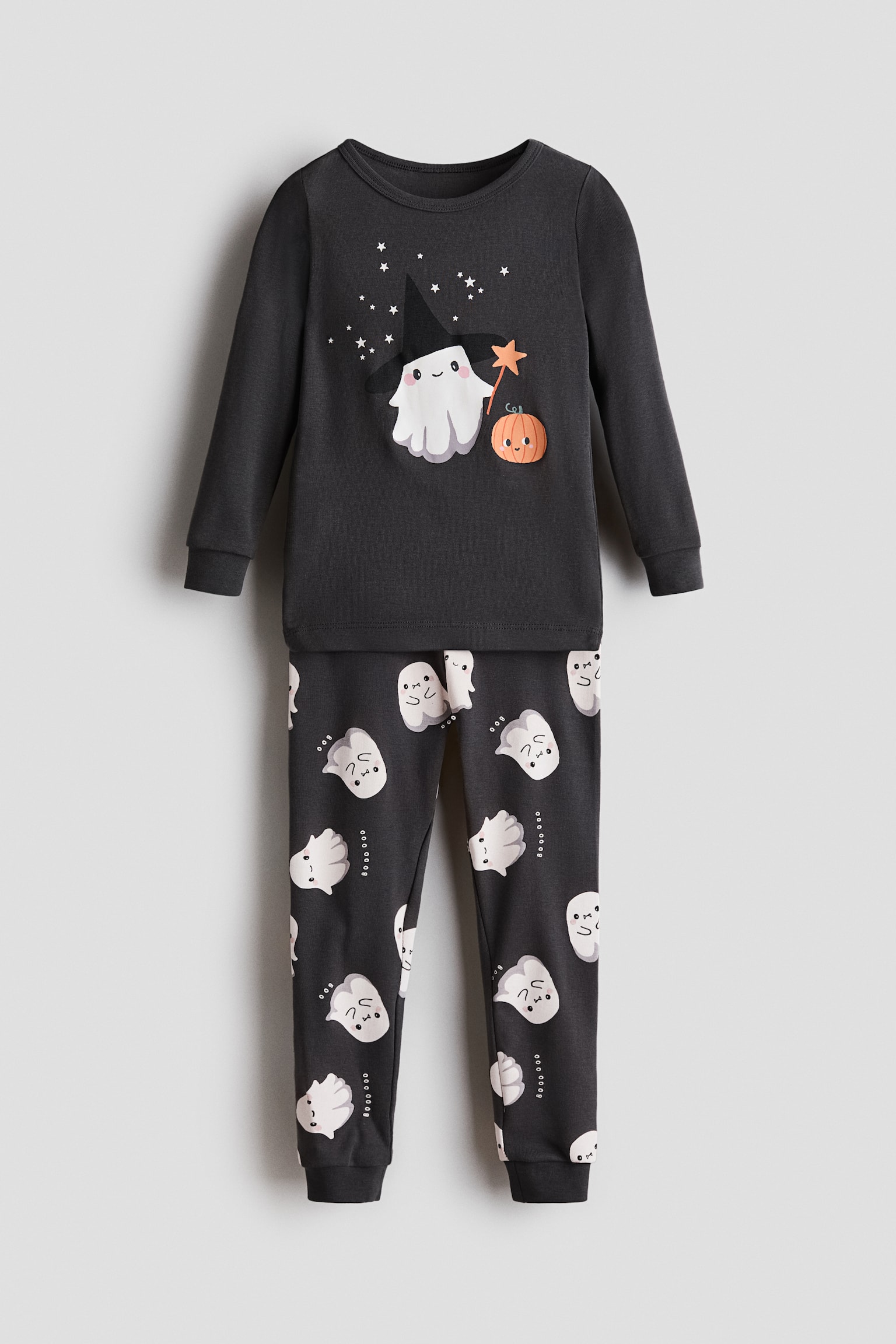 A children's two-piece pajama set in dark charcoal grey is shown from the front. The long-sleeve crewneck top is displaying a central graphic of a white ghost wearing a black witch hat and holding a star wand next to a small pumpkin, while the matching pants are featuring an all-over print of small white ghosts and ribbed ankle cuffs.