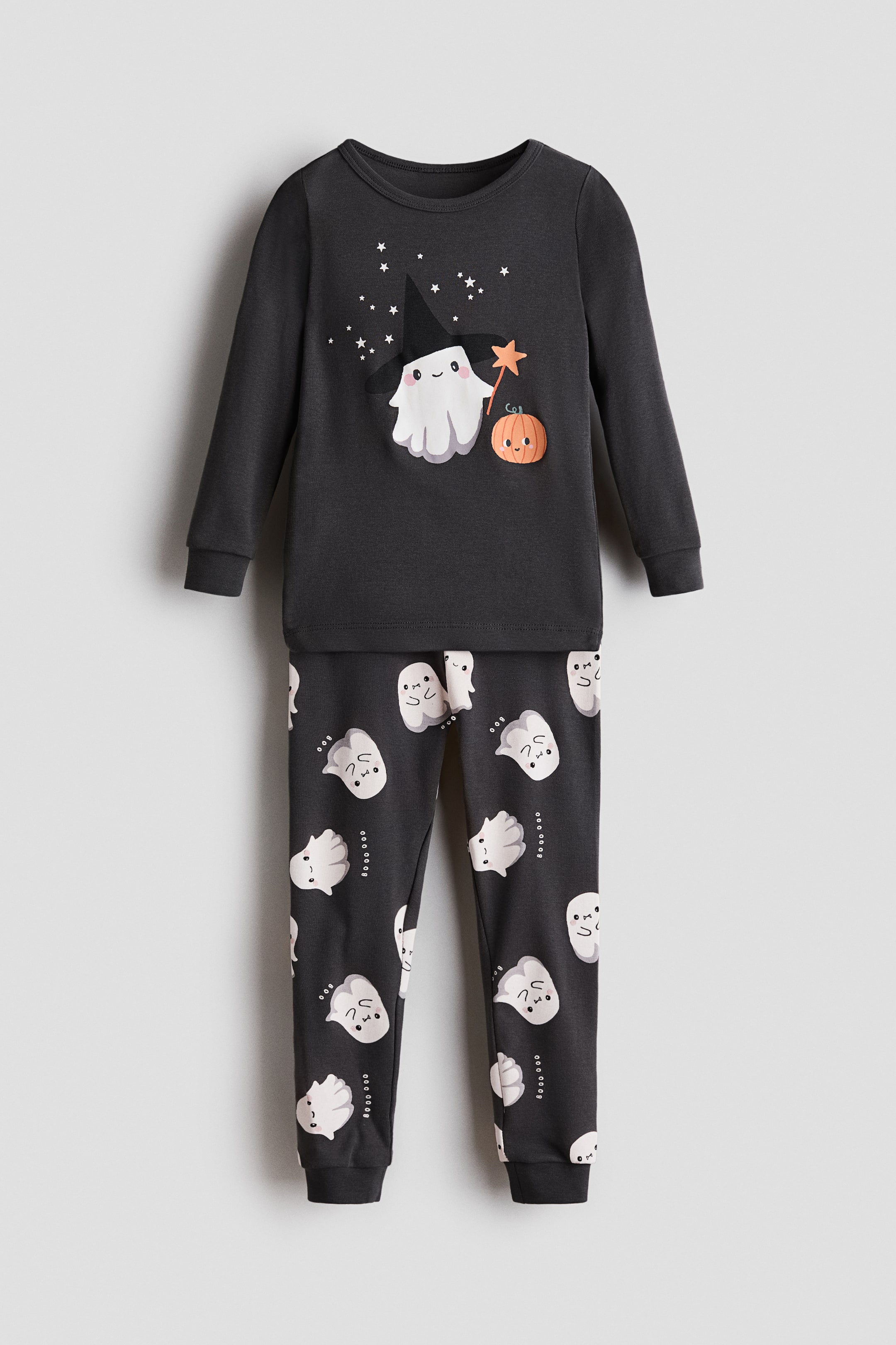 View larger image: A children’s two-piece pajama set in dark charcoal gray is shown from the front. The long-sleeve crewneck top features a central graphic of a white ghost wearing a black witch hat and holding a star wand next to a small pumpkin, while the matching pants feature an all-over print of small white ghosts and ribbed ankle cuffs.