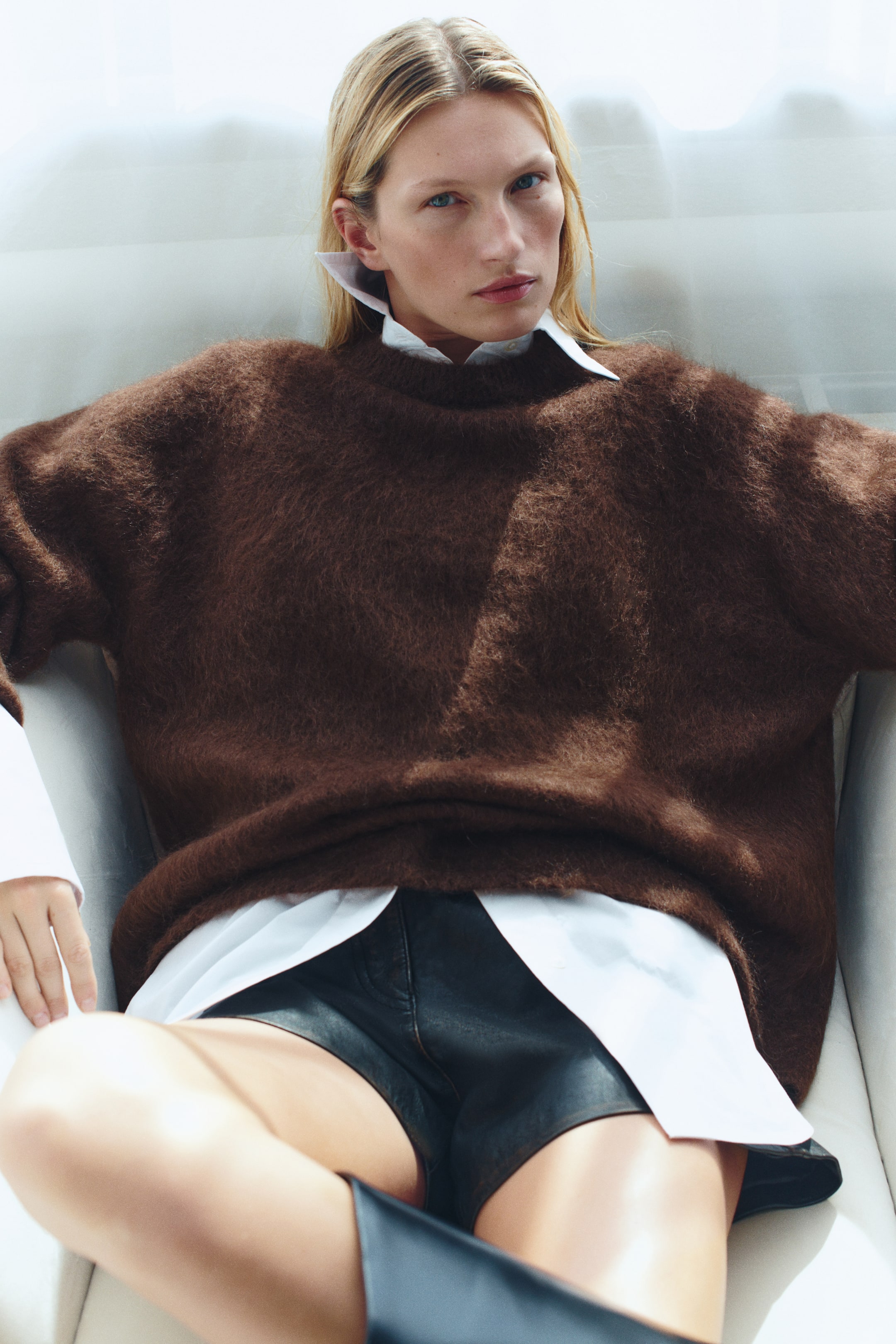Zobraziť väčší obrázok: Model má na sebe oversized hnedý sveter z mäkkého, hebkého úpletu, s padnutými ramenami a rebrovaným okrúhlym výstrihom. Sveter je vrstvený cez bielu košeľu s golierom a doplnený o čierne kožené šortky.